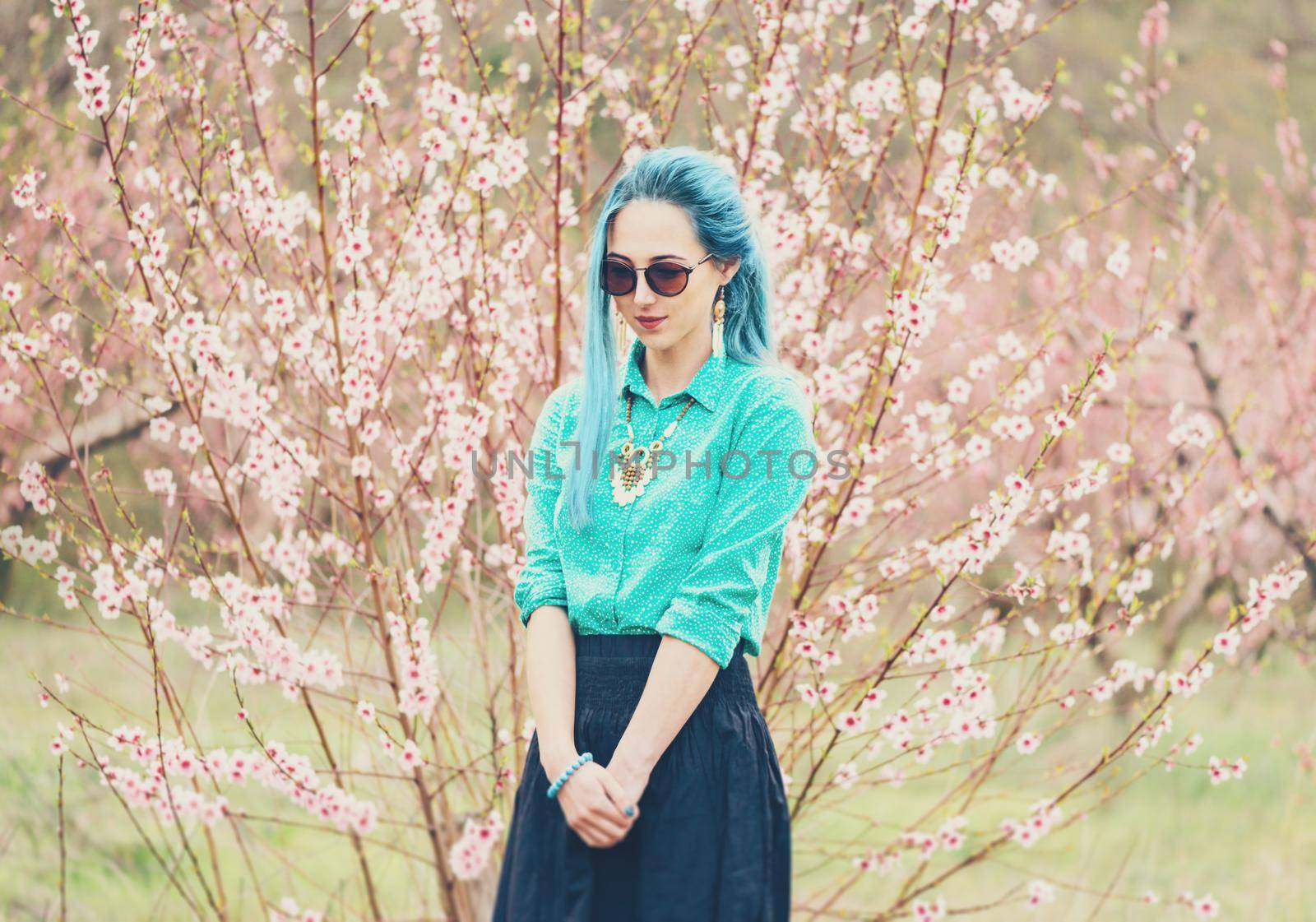 Attractive fashionable young woman with blue hair walking in blossoming peach tree garden in spring.