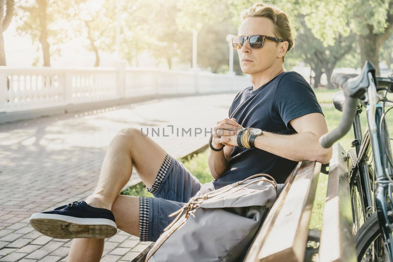 Handsome young man cyclist rests on wooden bench in summer park.
