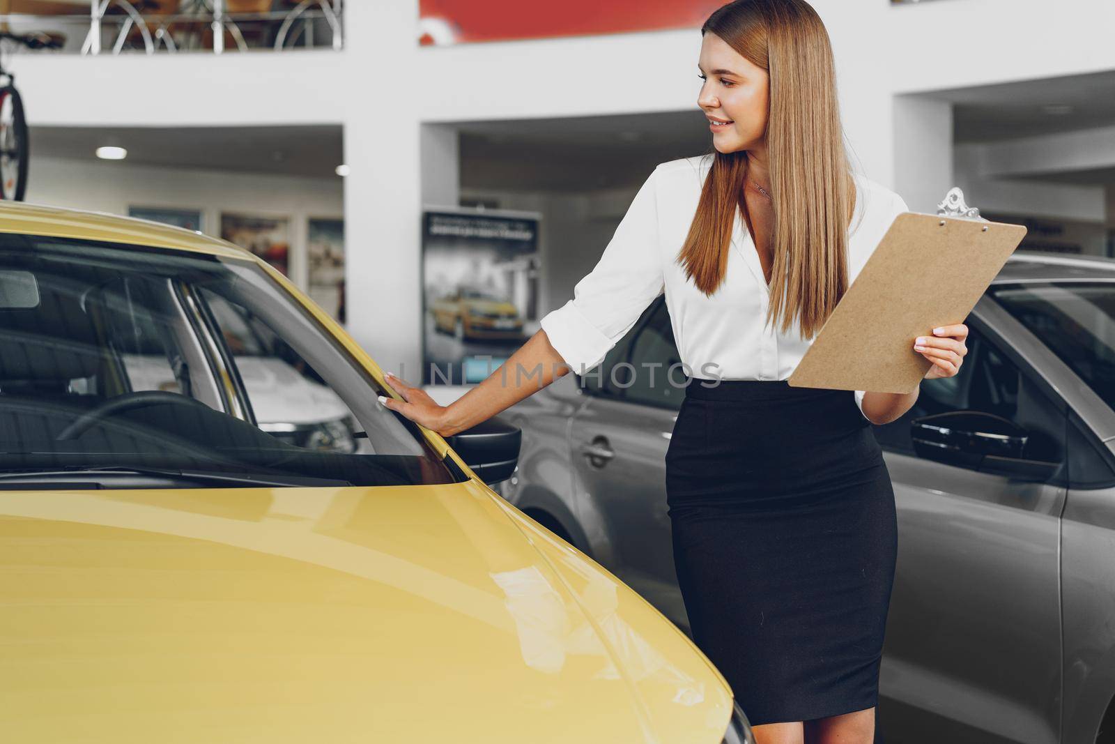 Attractive young female car dealer standing in showroom near a new car