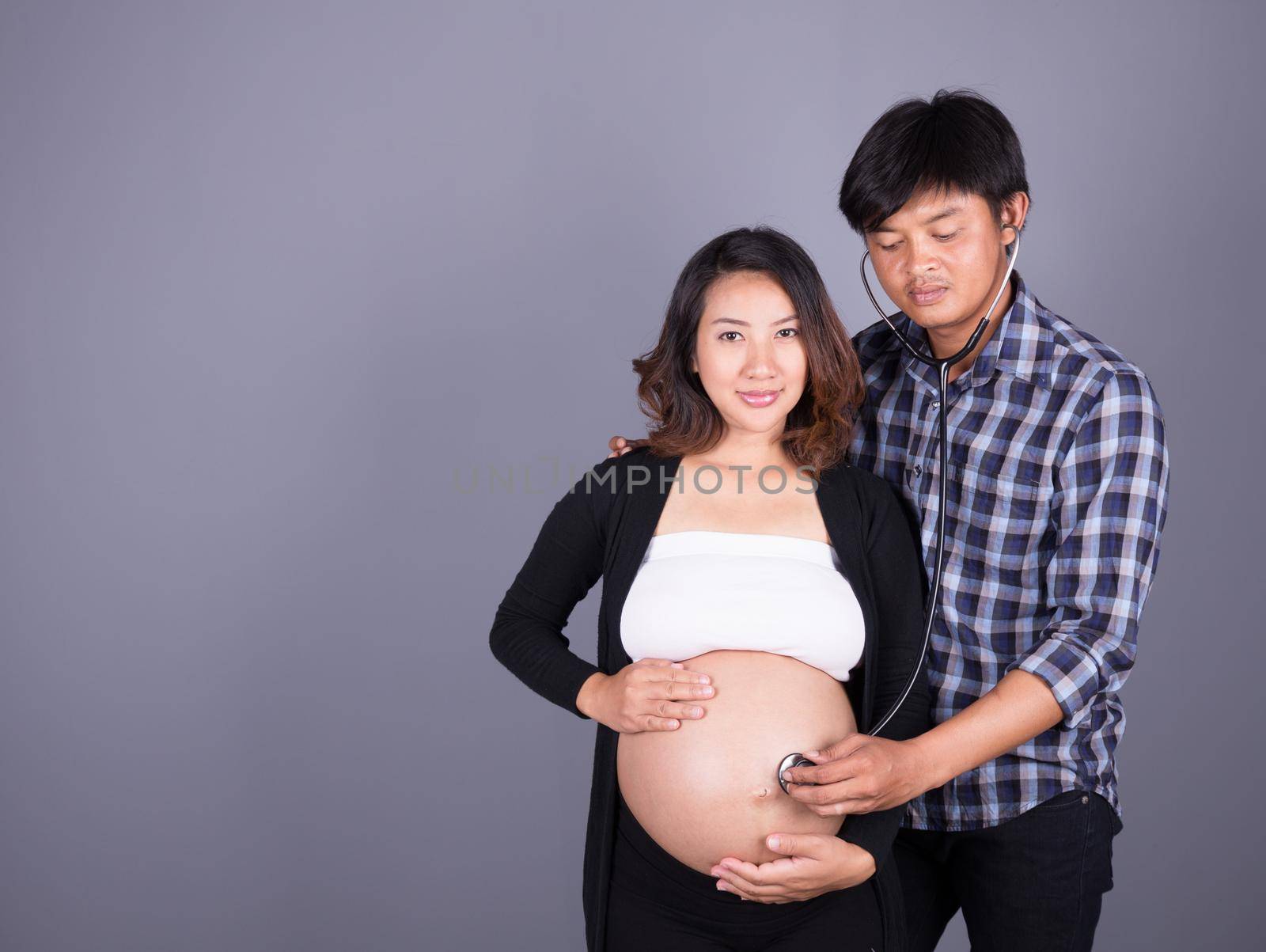 man listening the belly of his pregnant wife with stethoscope on gray background by geargodz