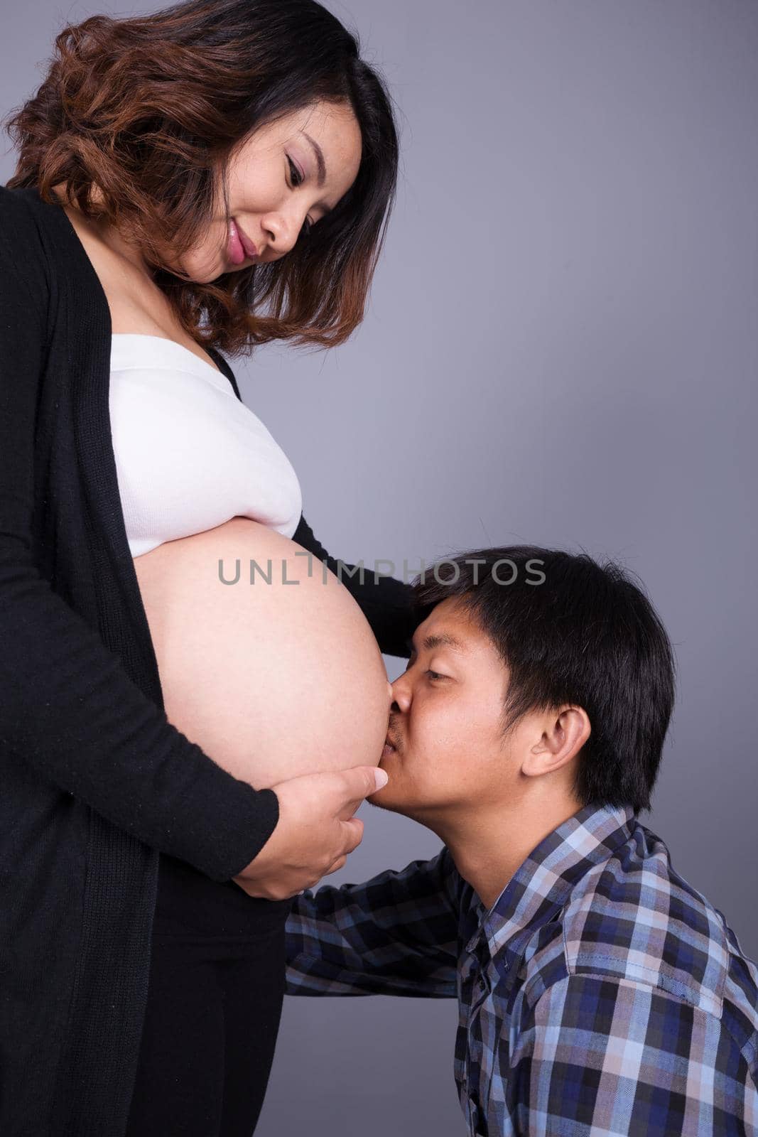 Young man kissing his pregnant wife's belly on gray wall background
