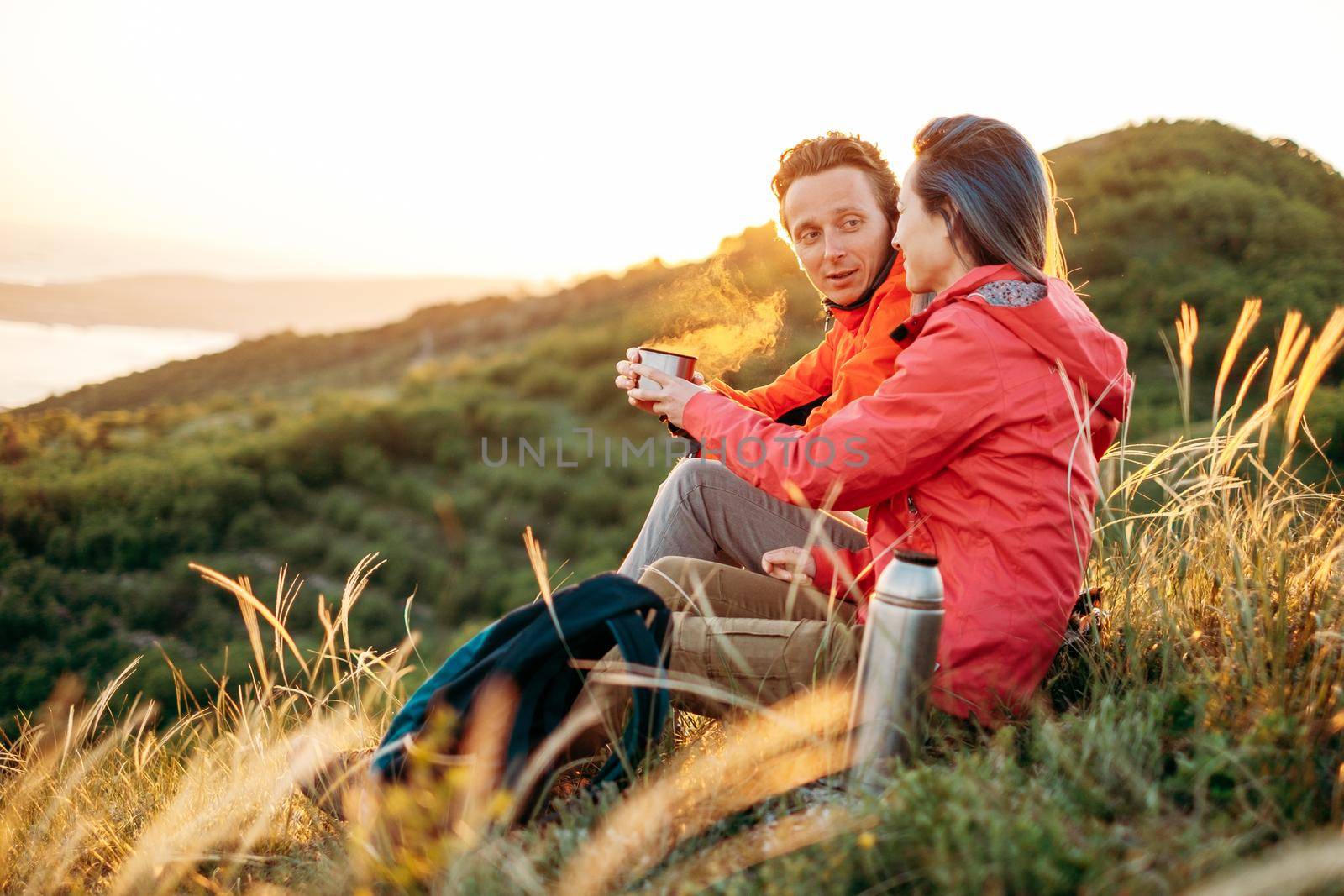 Traveler young couple resting in summer mountains at sunset. Man giving a cup of tea to woman.
