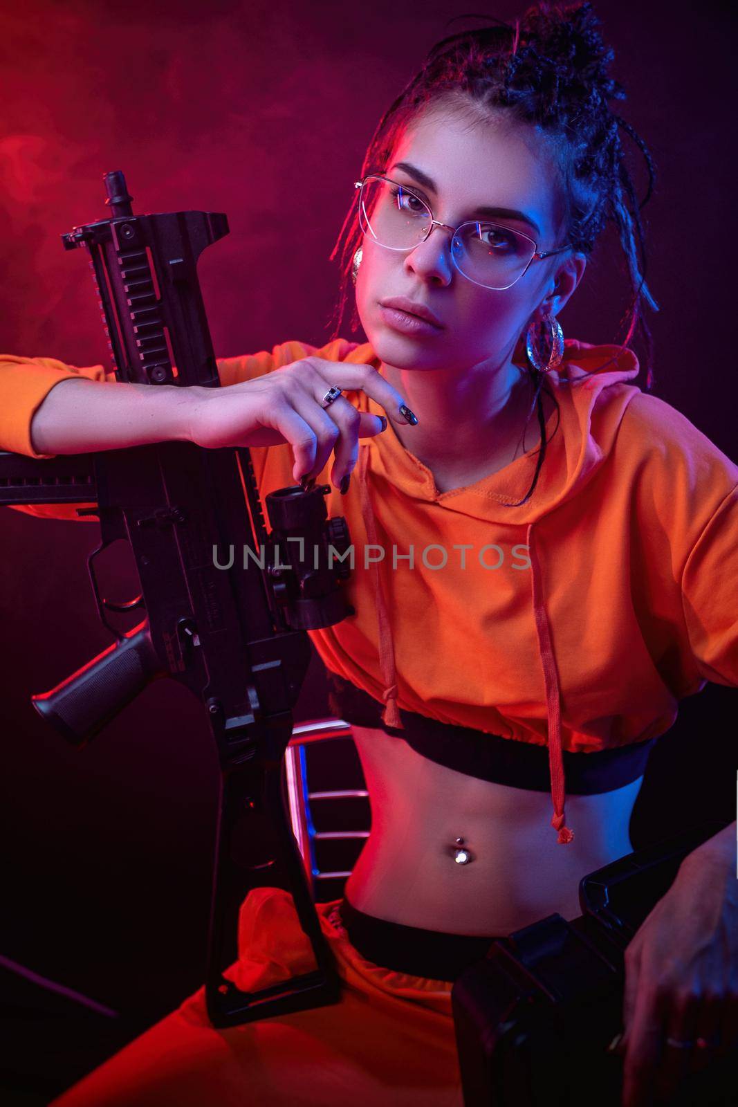 beautiful face brunette in a red jumpsuit with a short barrelled machine gun on a dark background in neon light