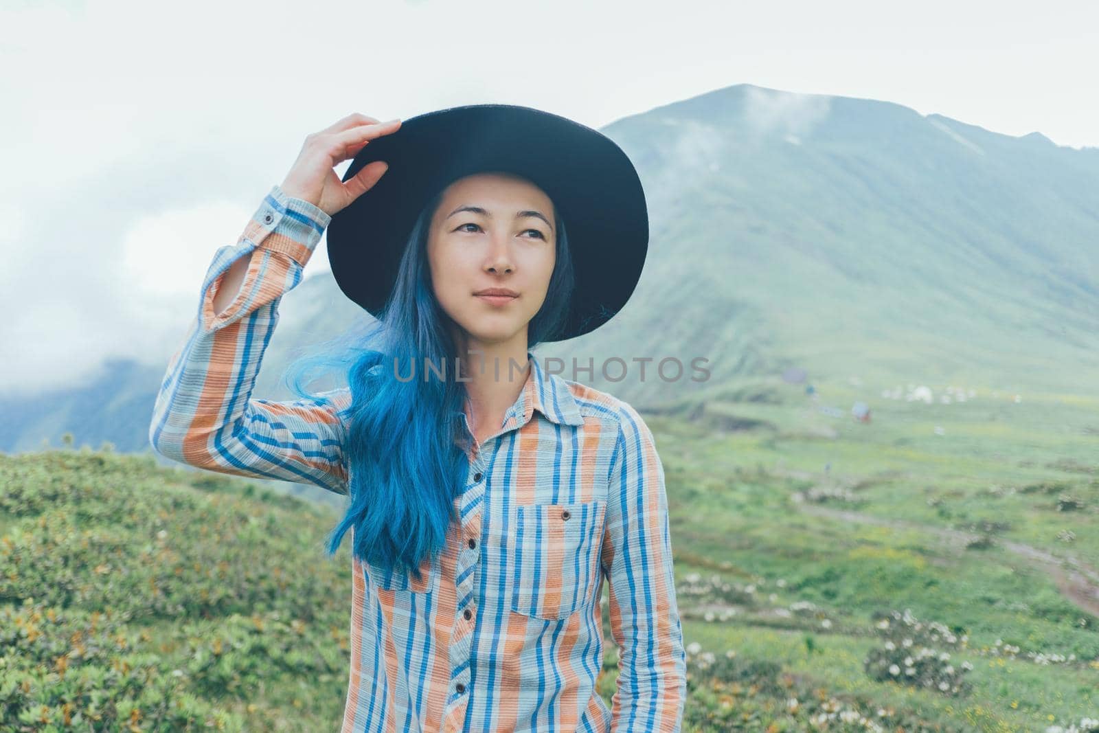 Beautiful young woman with blue hair wearing in a hat walking in summer mountains.