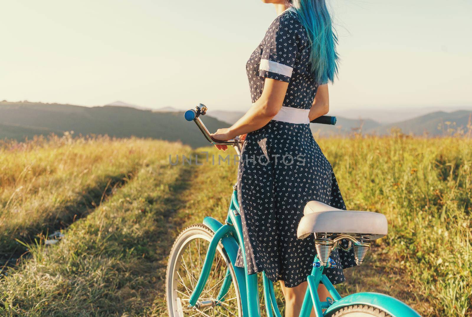 Unrecognizable young woman wearing in dress standing with bicycle cruiser on summer meadow.