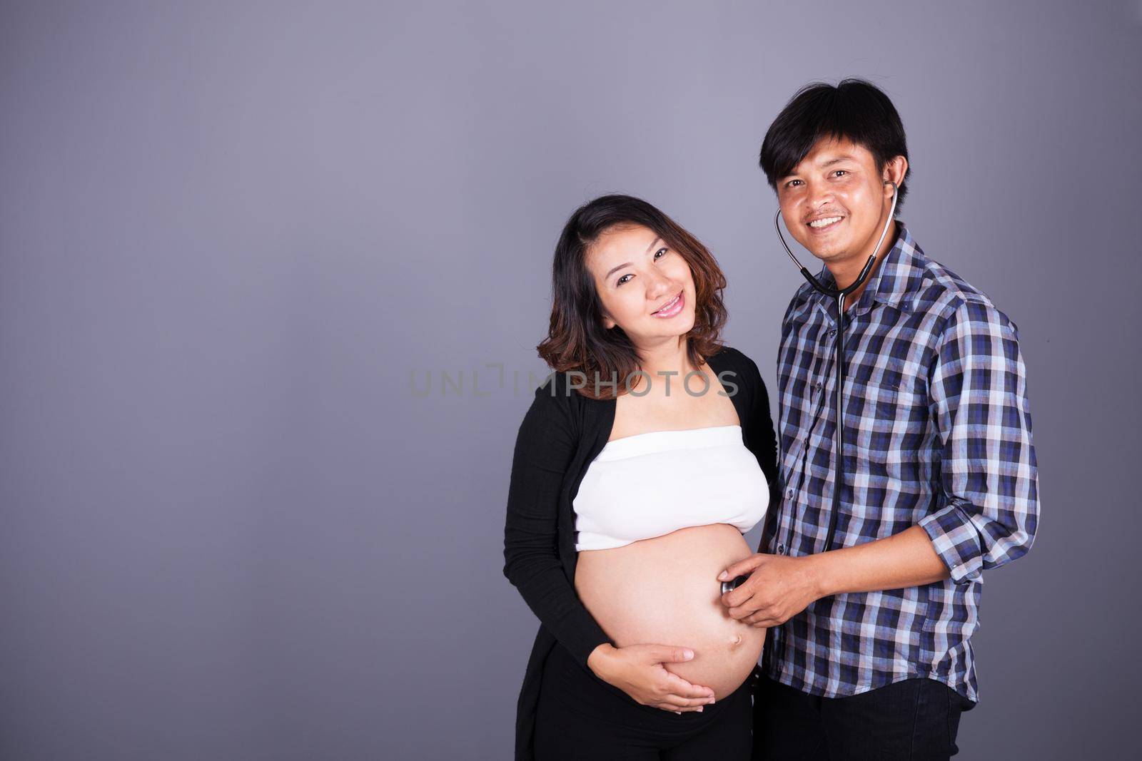 man listening the belly of his pregnant wife with stethoscope on gray background by geargodz
