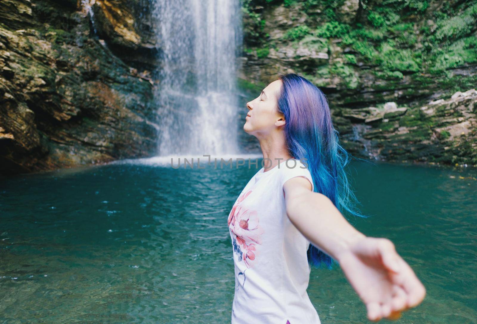 Freedom happy young woman standing with raised arms near the waterfall in summer, side view.