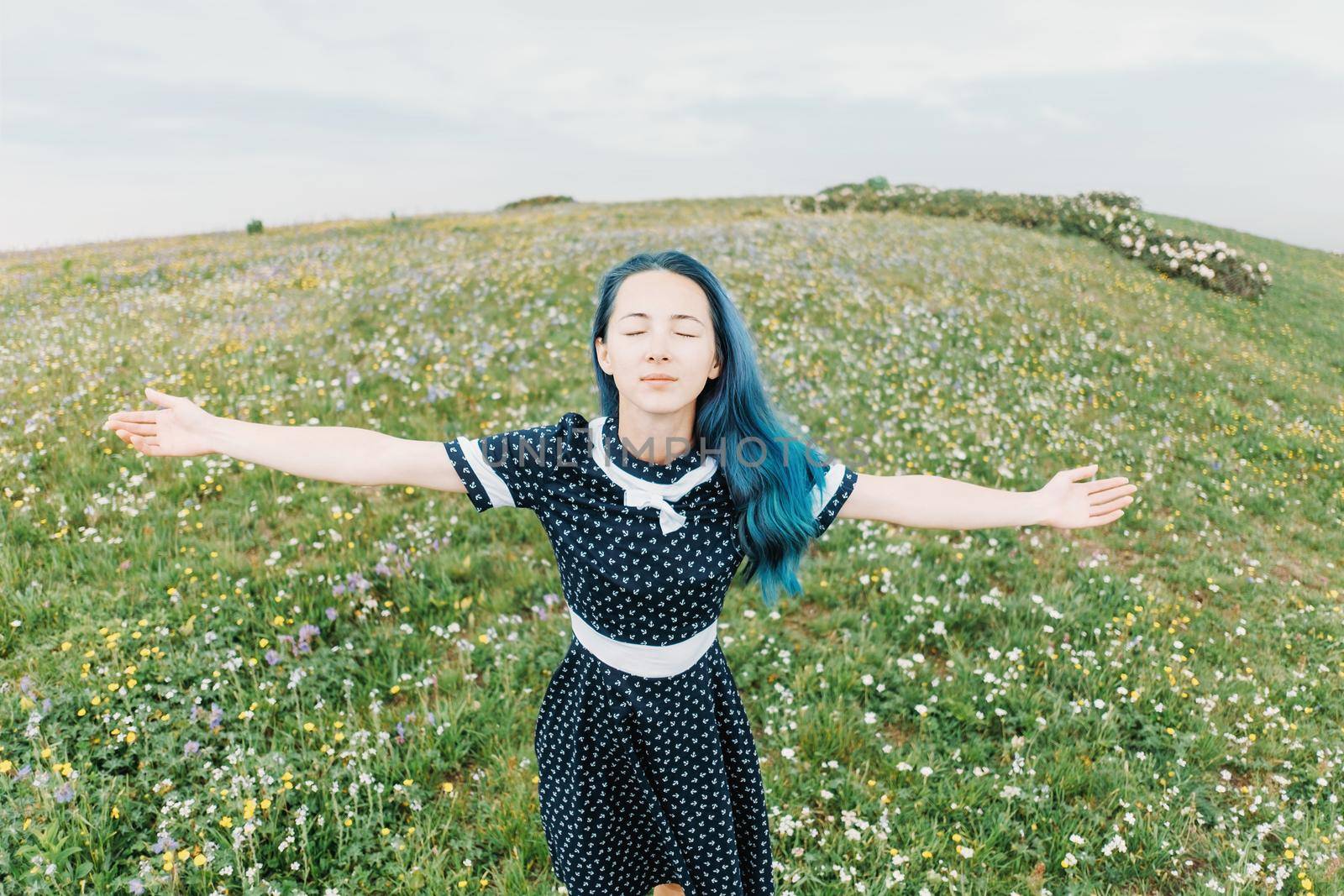 Beautiful young woman wearing in dress standing with raised arms on wildflower summer meadow, front view.