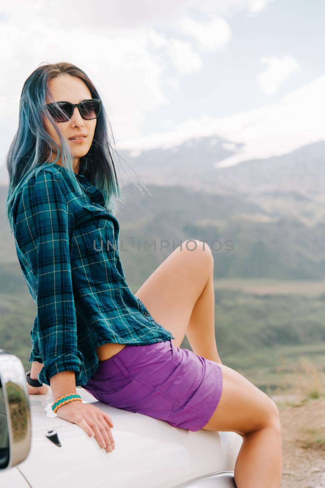 Explorer young woman sitting on car on background of mountains, looking at camera.