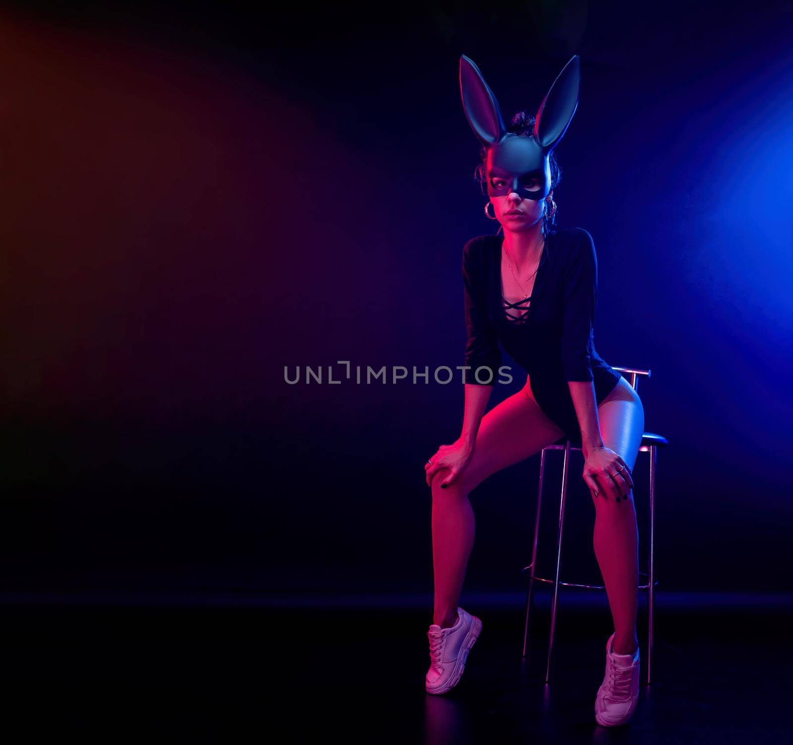 a beautiful girl in a bodysuit poses in a photo Studio on a dark background in neon light. On the face of a rabbit mask by Rotozey