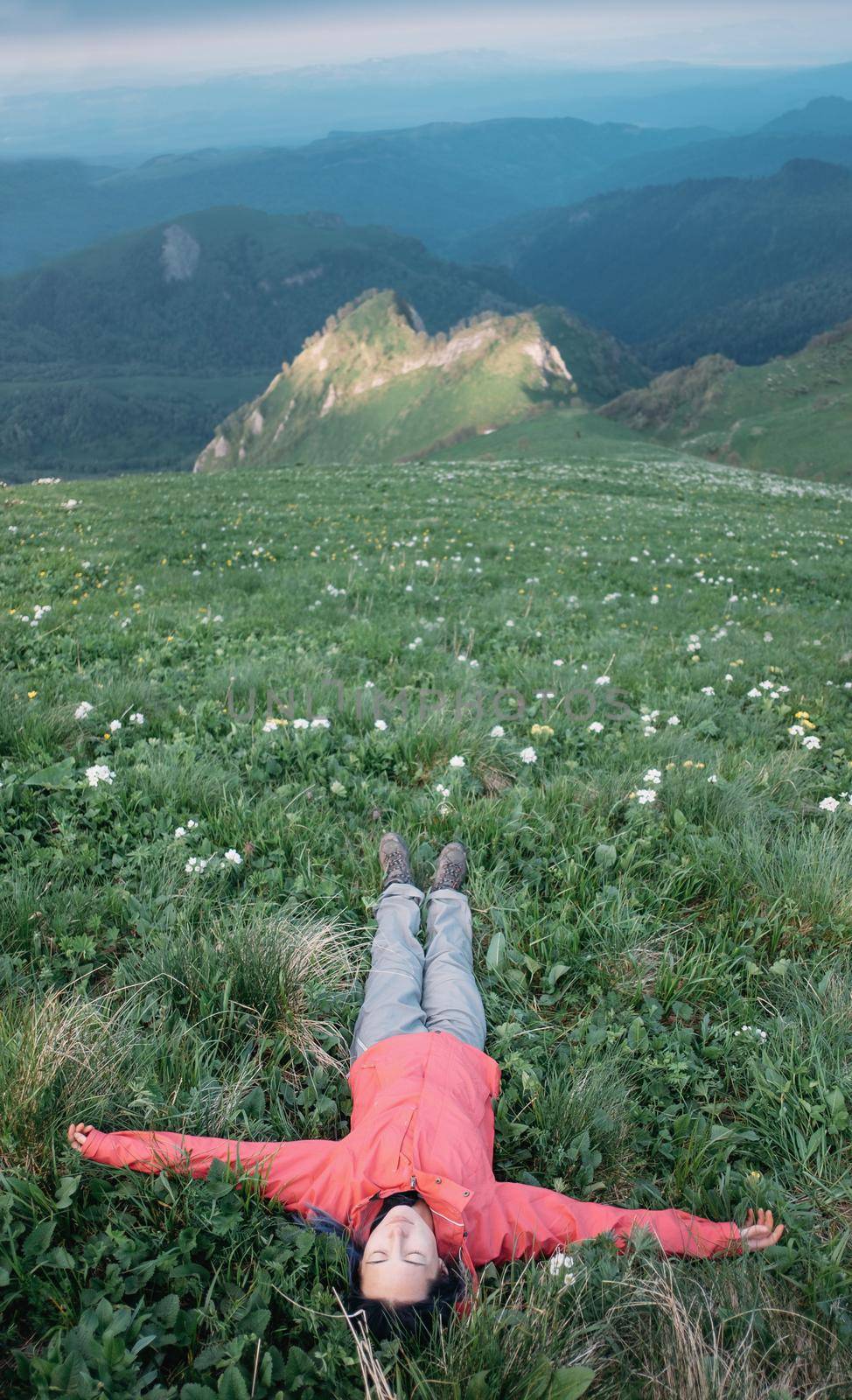 Happy traveler young woman lying with raised arms on wildflower meadow in summer mountains.