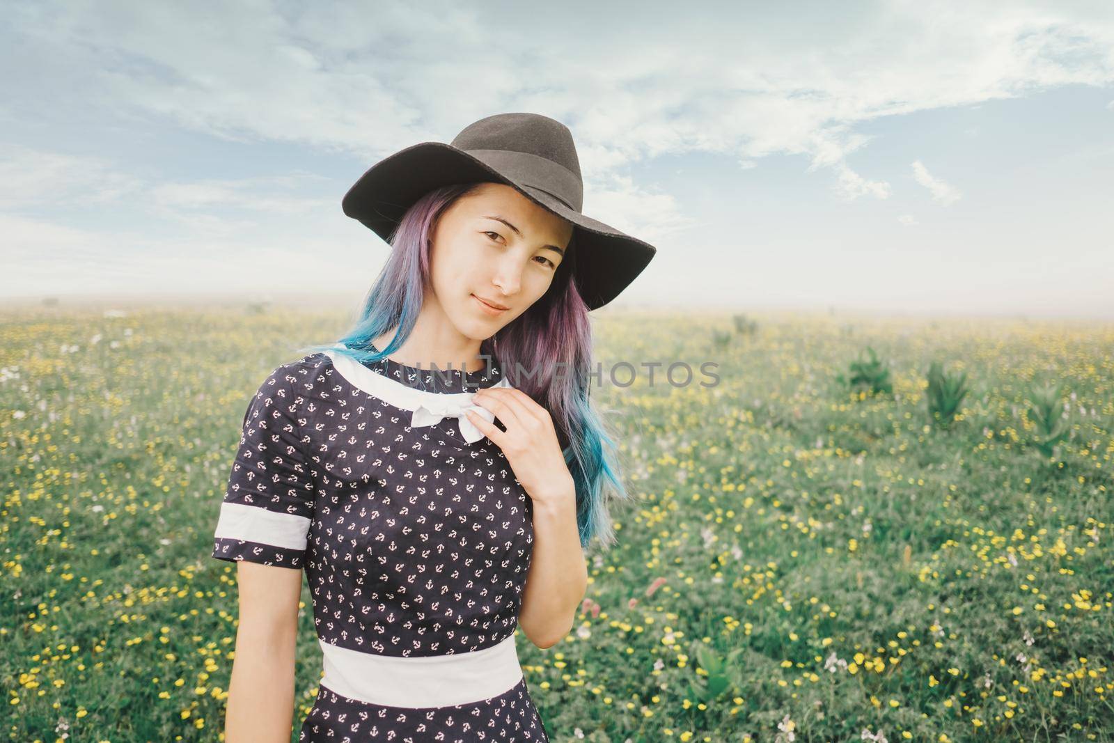 Beautiful young woman wearing in dress and hat walking on wildflower meadow, looking at camera.