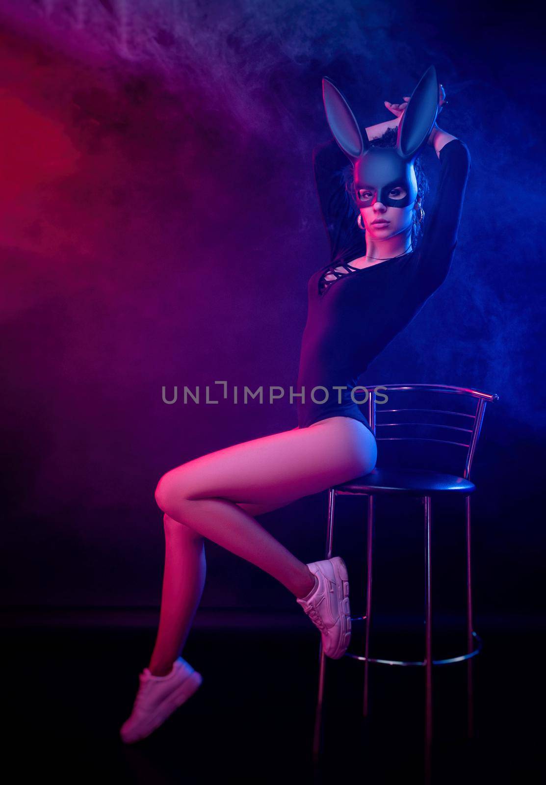 a beautiful girl in a bodysuit poses in a photo Studio on a dark background in neon light. On the face of a rabbit mask by Rotozey