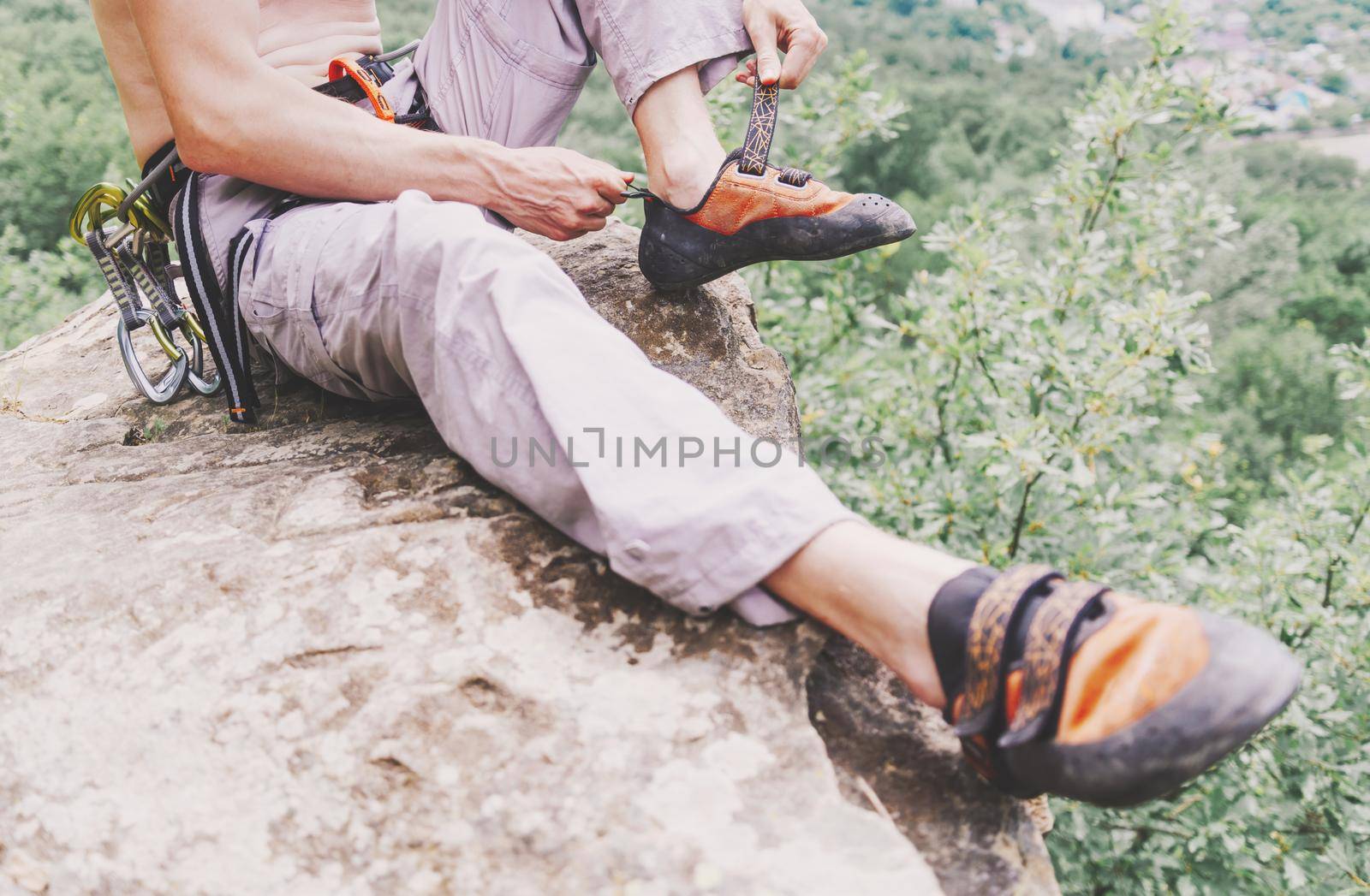 Sporty young man sitting on cliff and putting on climbing shoes in summer.