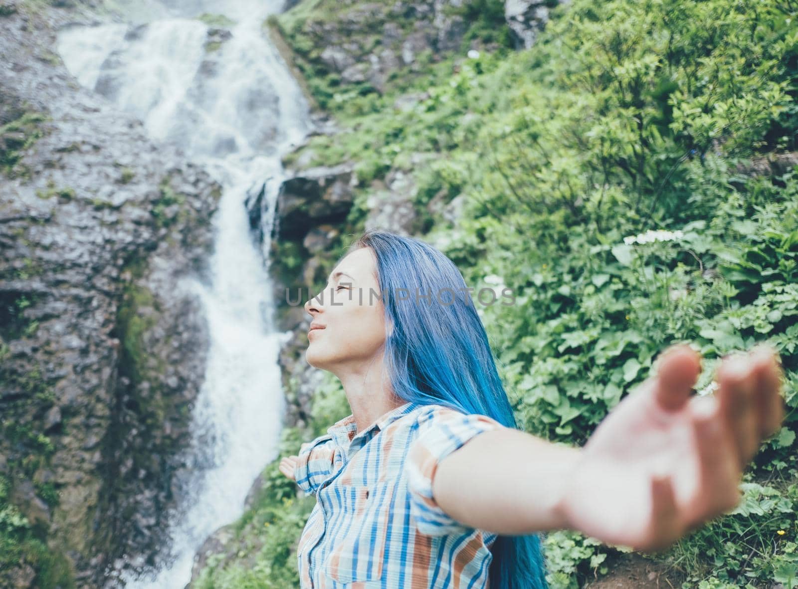 Happy young woman standing with raised arms on background of waterfall and enjoying of summer nature.