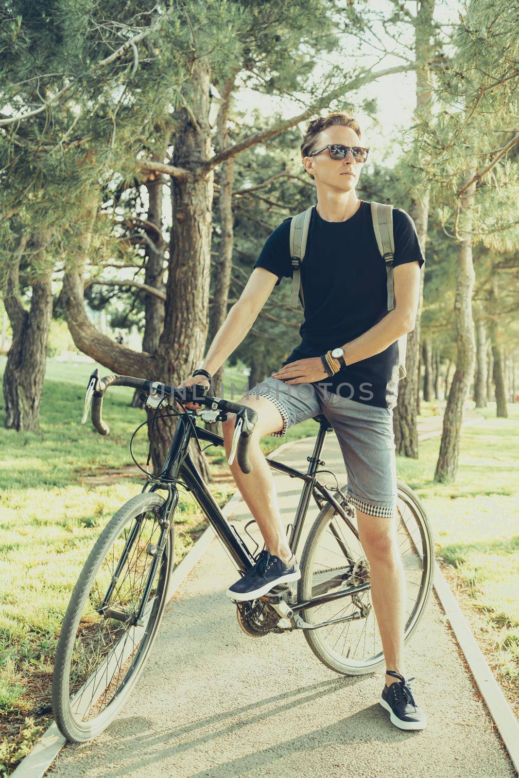 Handsome young man in sunglasses standing with bike in summer park outdoor.