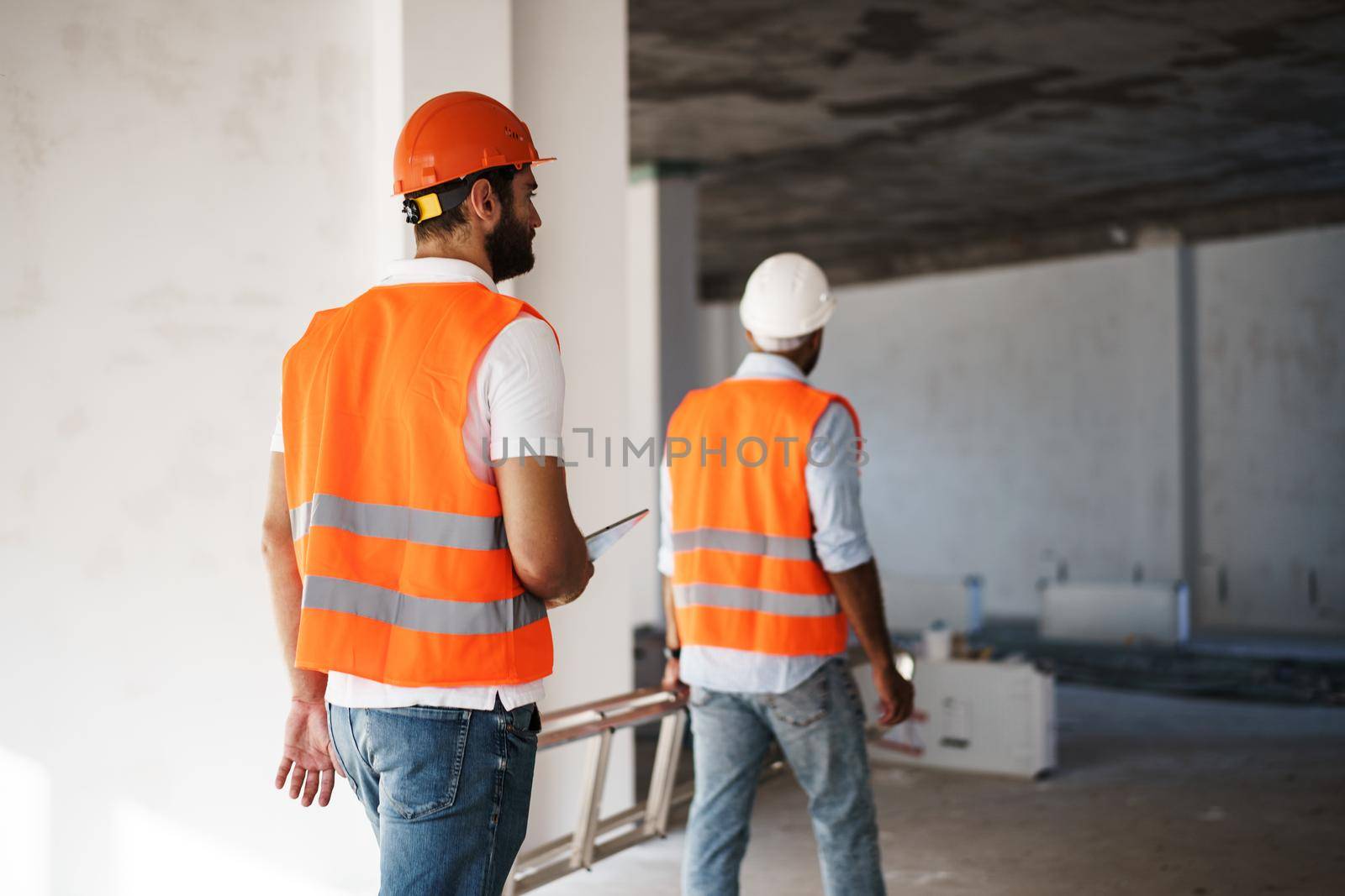 Two young male engineers in uniform and hardhats working at construction site, close up