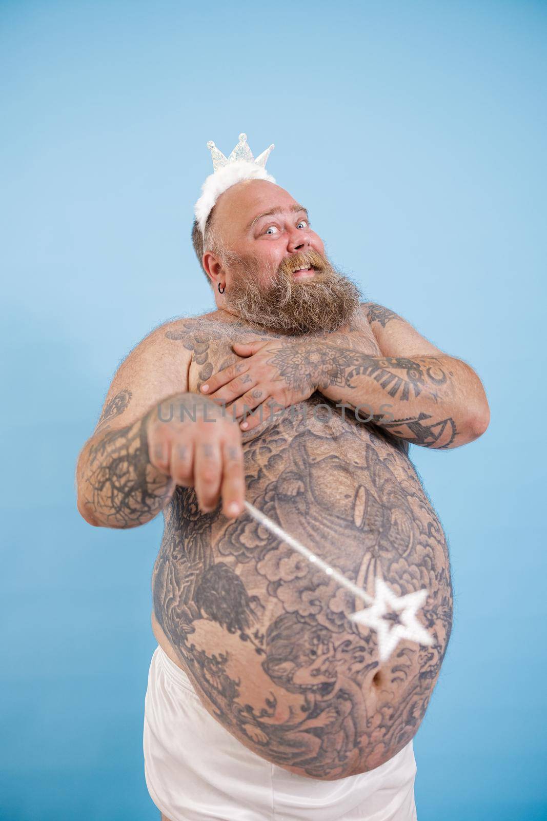 Smiling mature plus size man with toy crown and magic stick covers breasts by hands posing on light blue background in studio