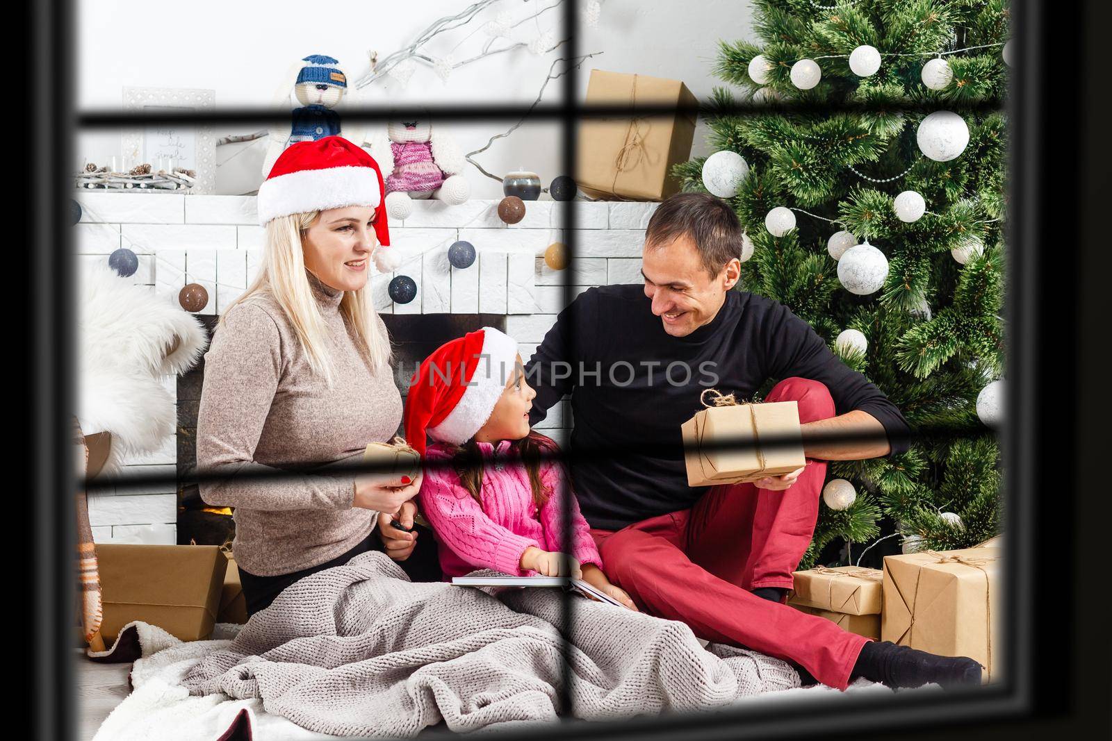 Young big family celebrating Christmas enjoying dinner, view from outside through a window into a decorated living room with tree and candle lights, happy parents eating with three kids