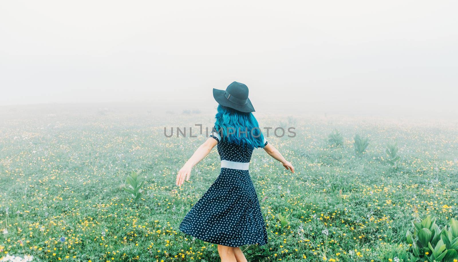Young woman wearing in dress and hat walking on wildflower summer meadow in foggy weather.