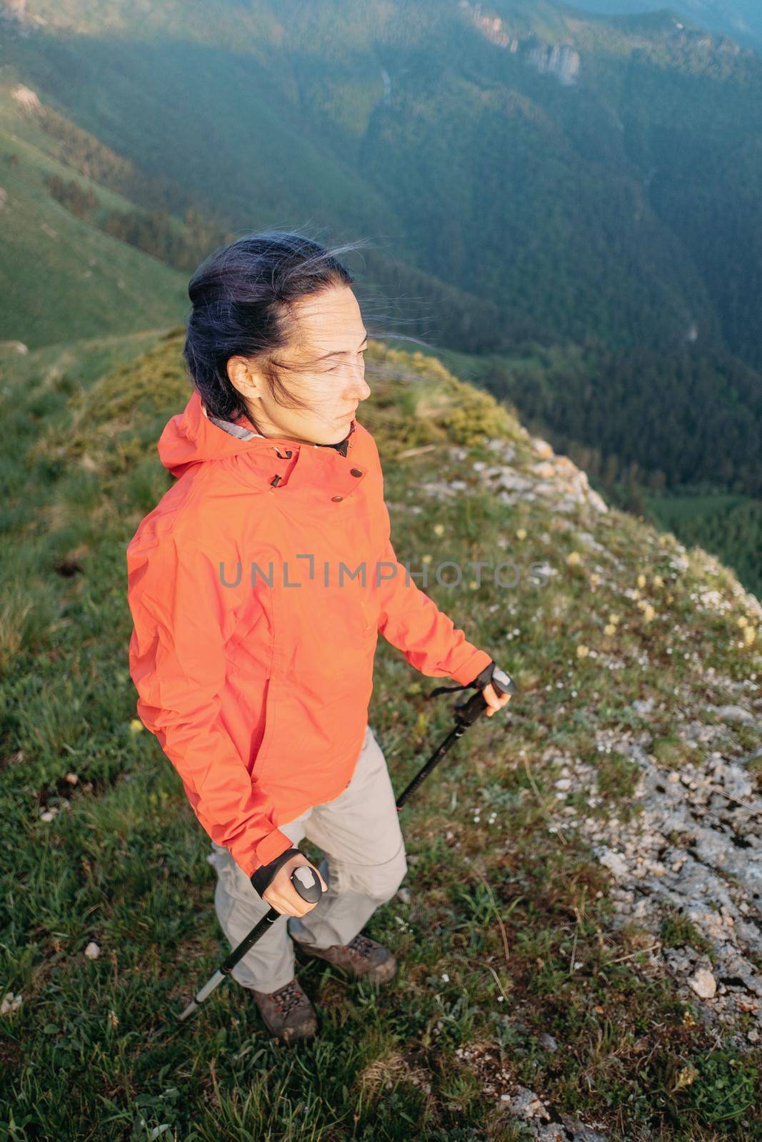 Hiker young woman walking with trekking poles in summer mountains in windy weather.