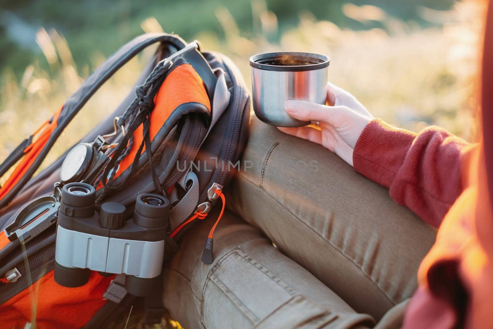 Unrecognizable explorer young woman rests with cup of tea and backpack in summer.