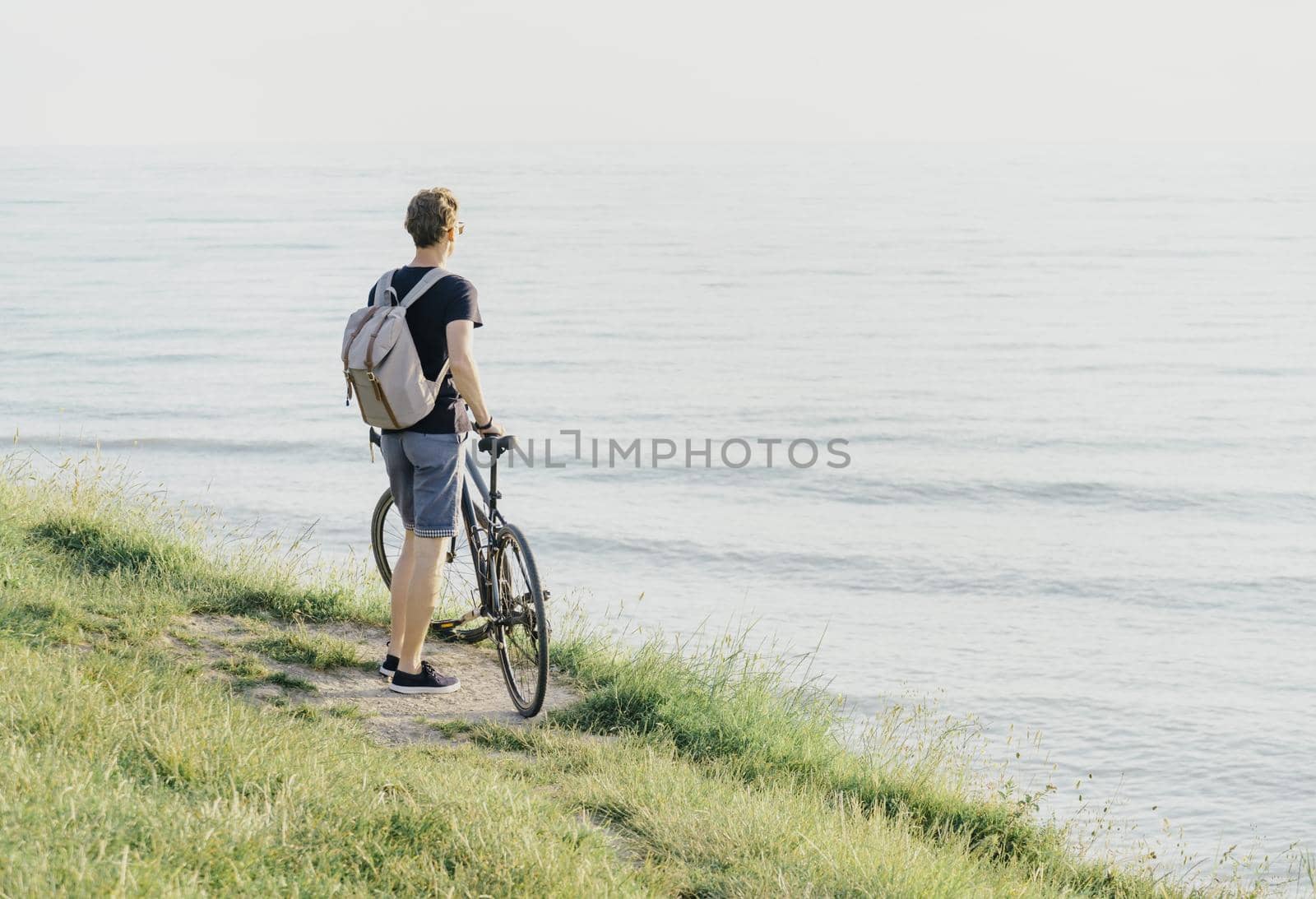 Young man with backpack and bicycle standing on coast and enjoying view of sea.