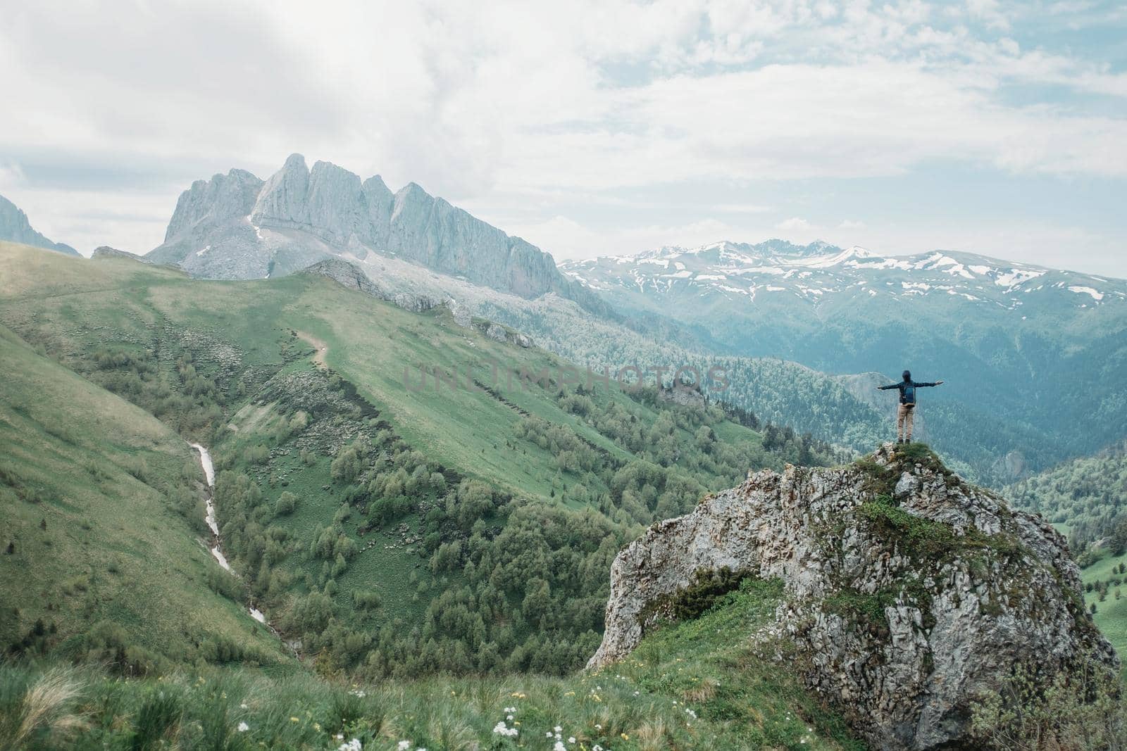 Hiker standing on peak of cliff with raised arms on background of summer mountains. Small figure of explorer on nature.