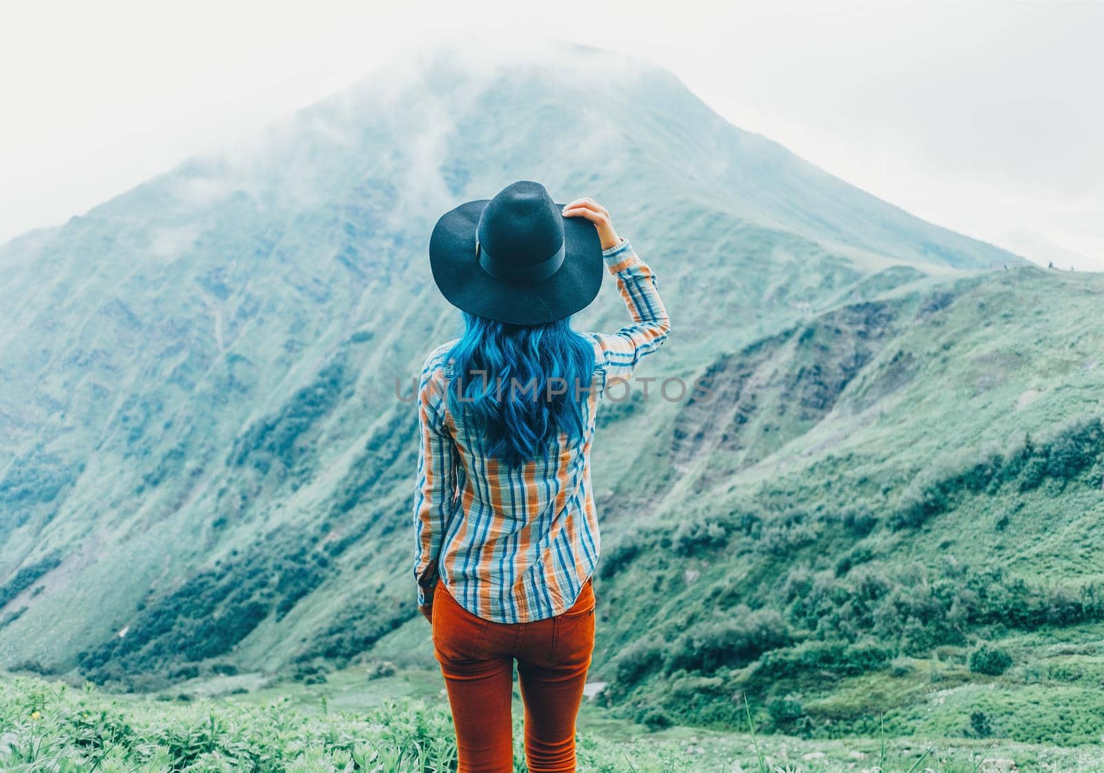 Explorer young woman in a hat standing in front of mountains in summer, rear view.