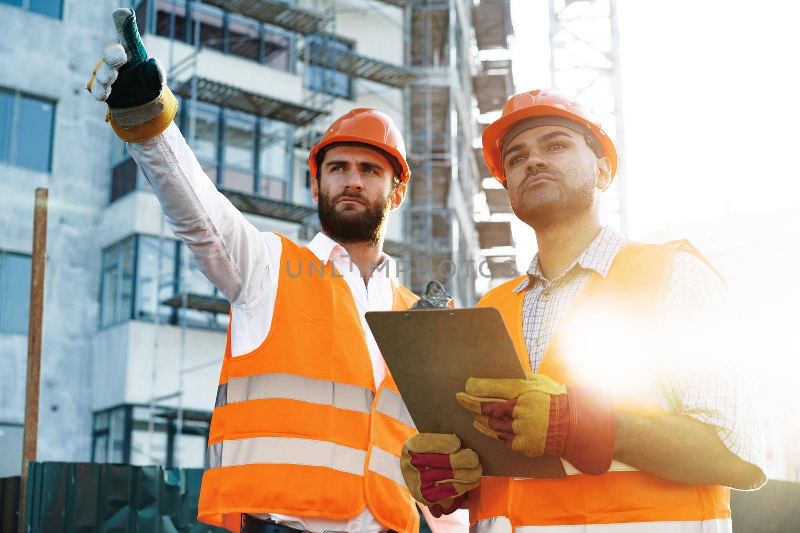 Two young male engineers in uniform and hardhats working at construction site, close up