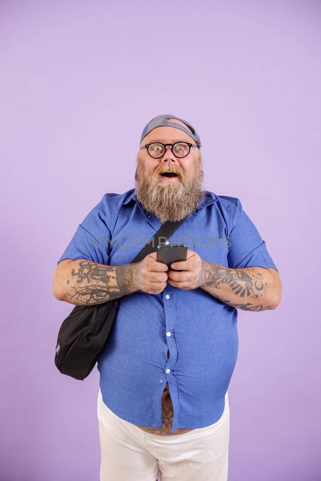 Funny surprised plus size man with crossbody bag and glasses holds mobile phone standing on purple background in studio