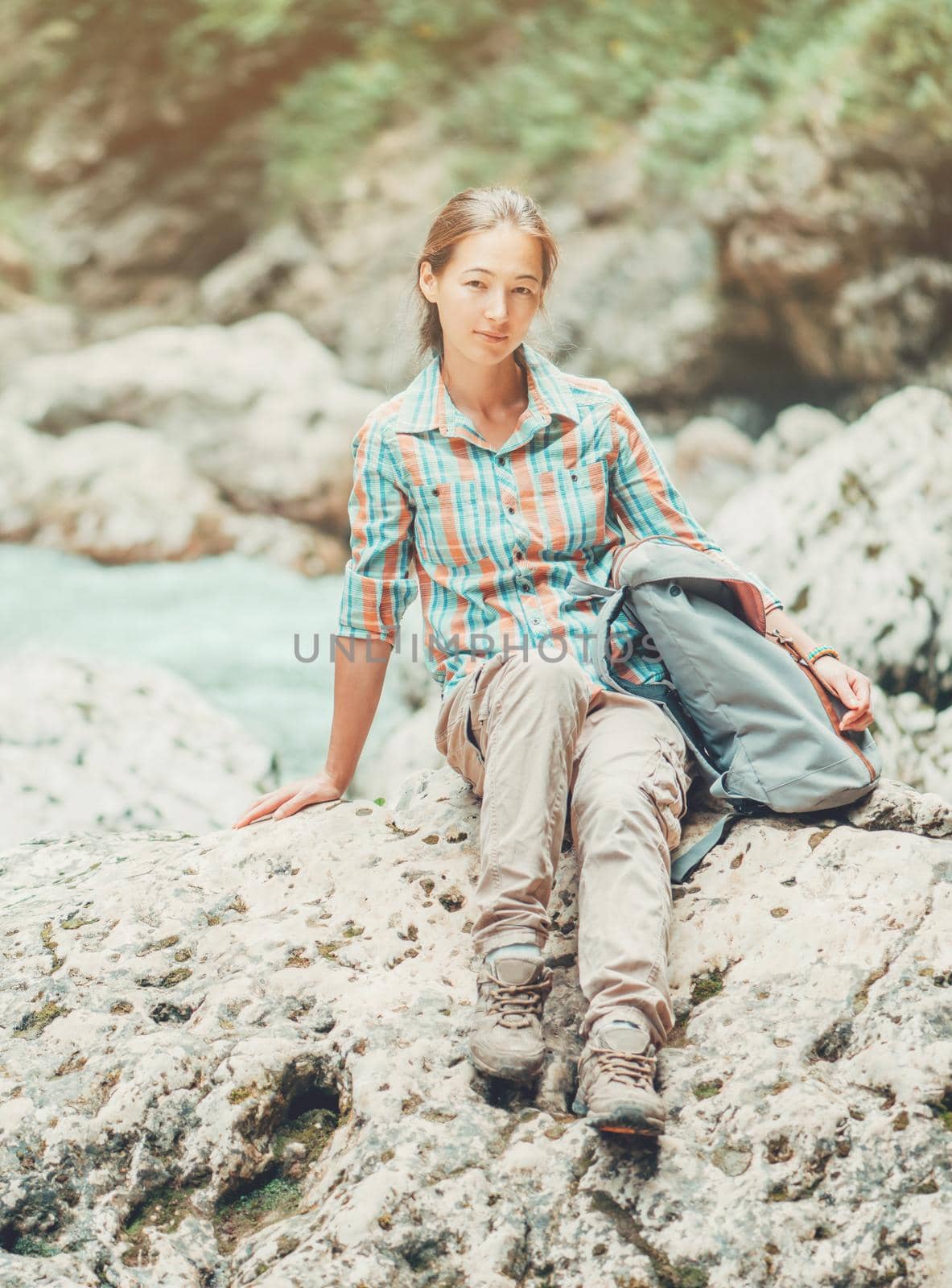 Explorer young woman wearing in cargo pants and plaid shirt sitting on rocky stone in summer, looking at camera.
