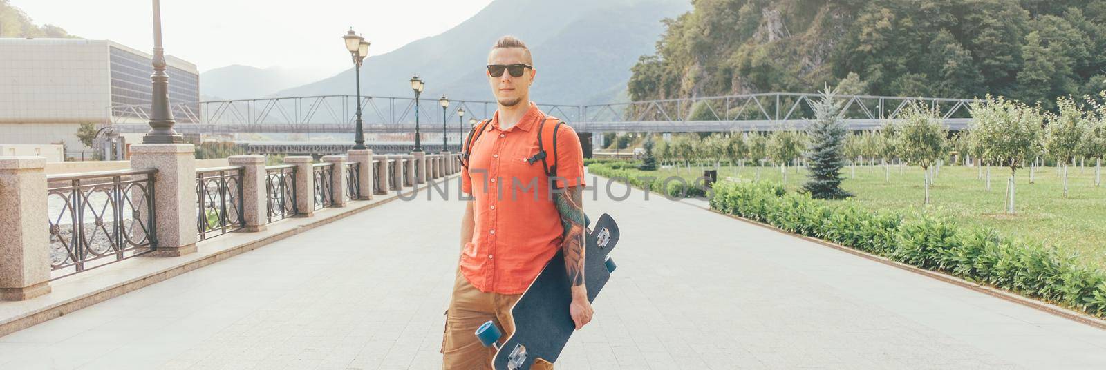 Street style tattooed guy in sunglasses walking with longboard in summer park.