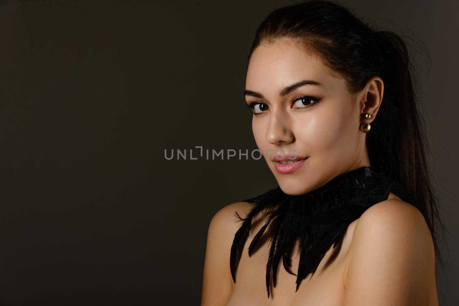 Portrait of a beautiful brunette girl with hair gathered in a ponytail with an ornament on the neck of the black feathers on dark background