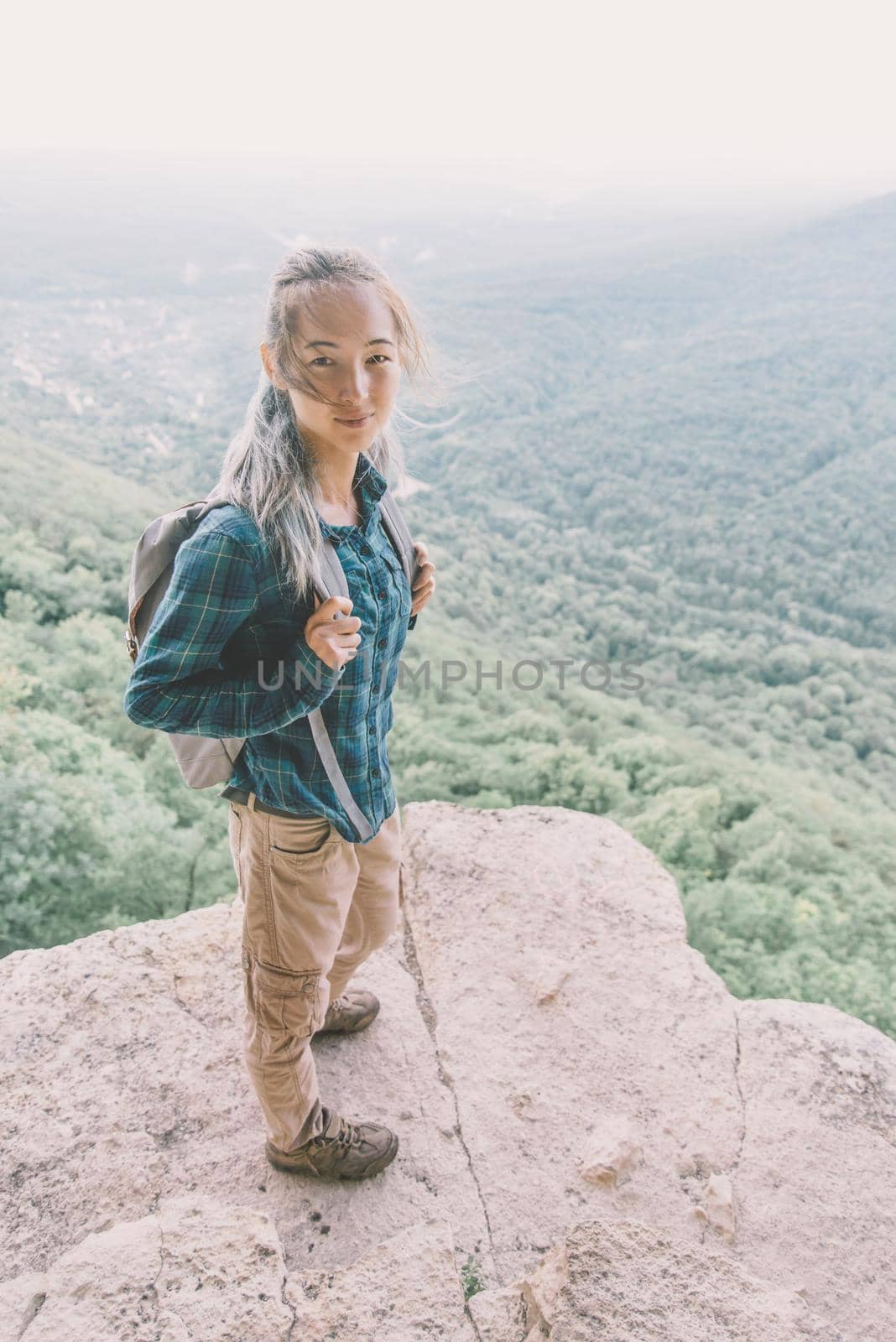Traveler young woman with backpack standing on high cliff over summer valley, looking at camera.
