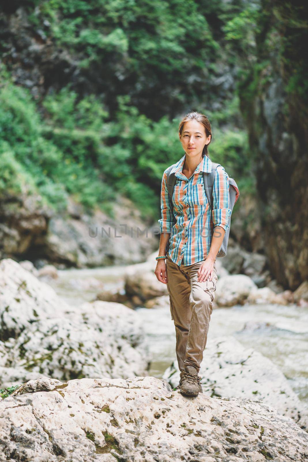 Explorer young woman wearing in cargo pants and plaid shirt standing on rocky stone outdoor, looking at camera.