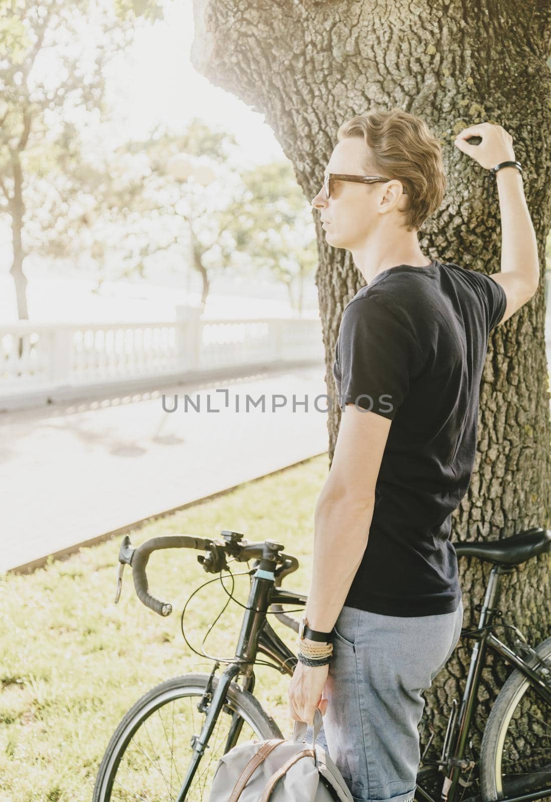 Young man standing with bicycle near a tree in summer park.