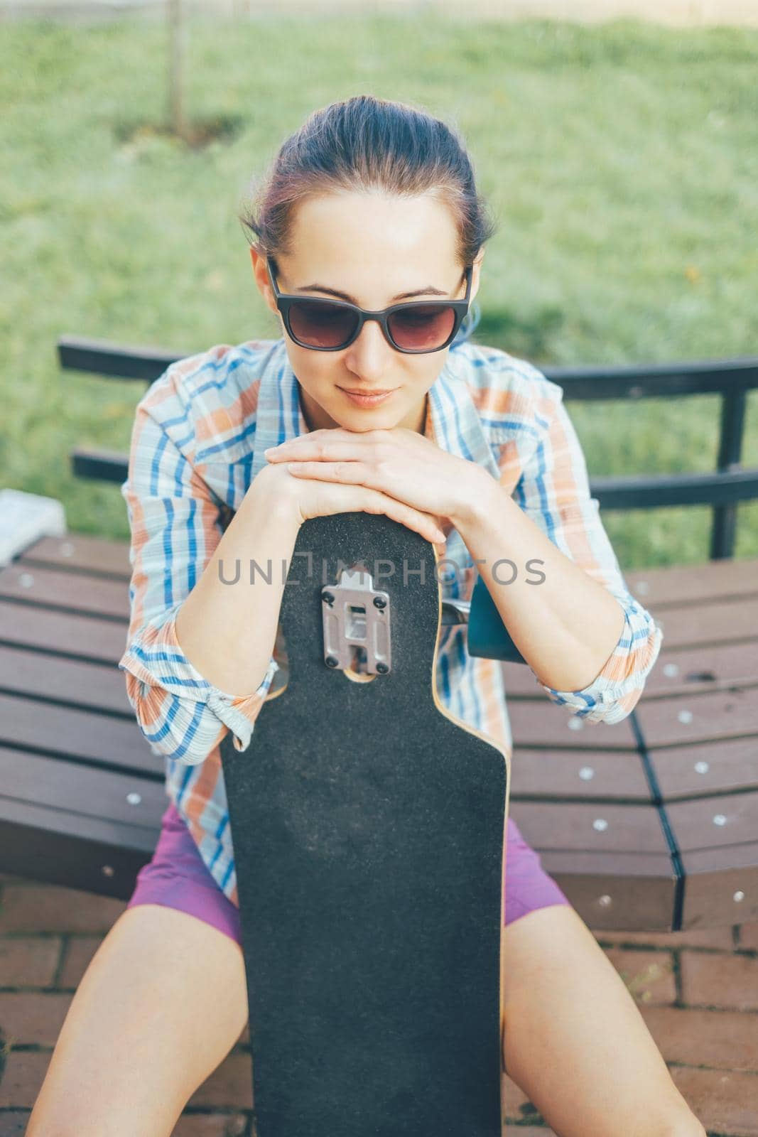 Young woman in sunglasses sitting with longboard on wooden bench outdoor.