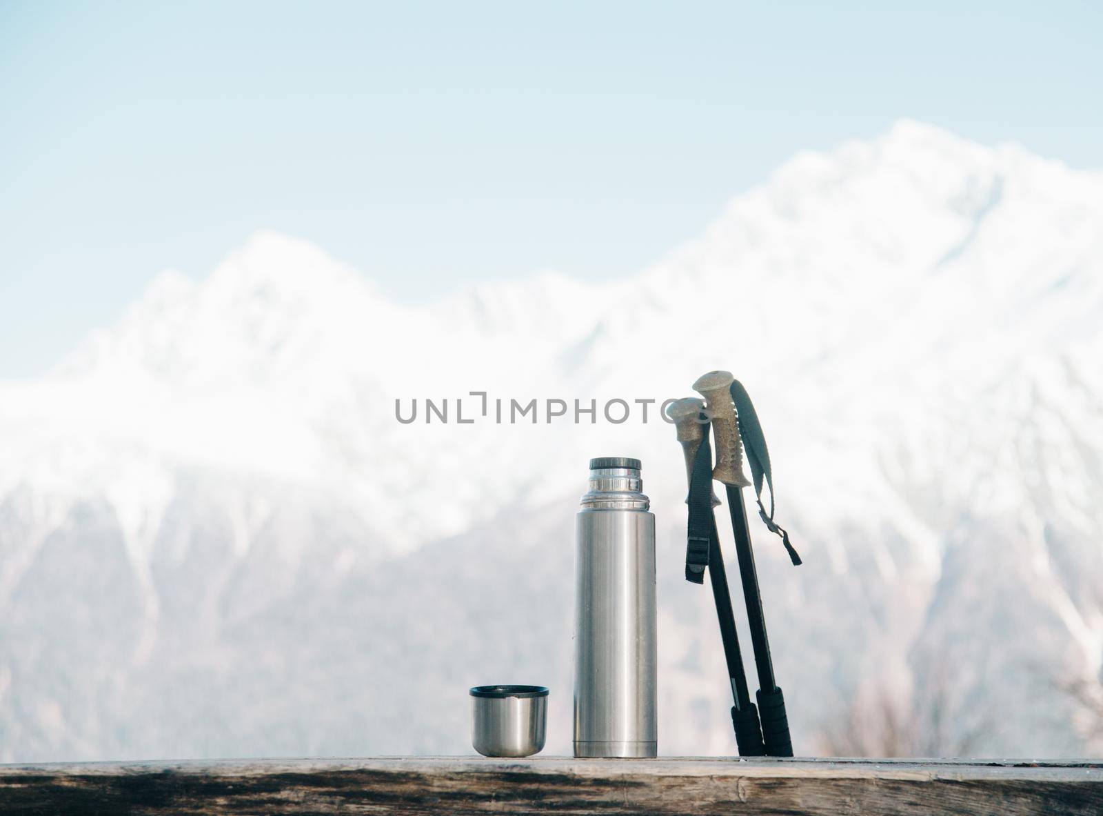 Thermos and trekking poles on wooden table on background of snowy mountains in winter.