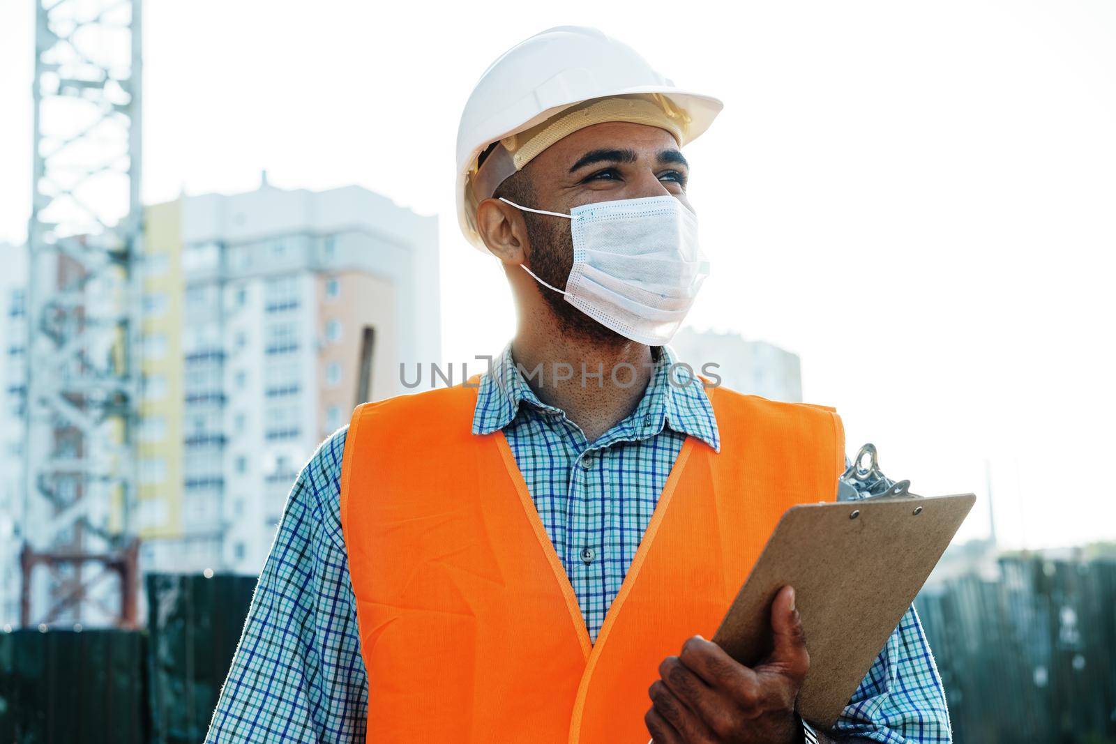 Portrait of mixed race man builder in workwear and hardhat wearing medical mask, close up photo