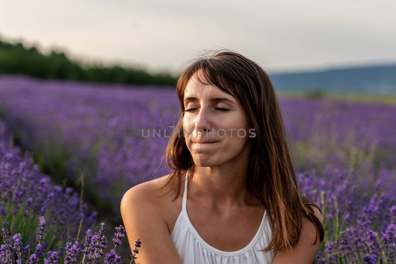 Close up portrait of happy young brunette woman in white dress on blooming fragrant lavender fields with endless rows. Warm sunset light. Bushes of lavender purple aromatic flowers on lavender fields. by panophotograph