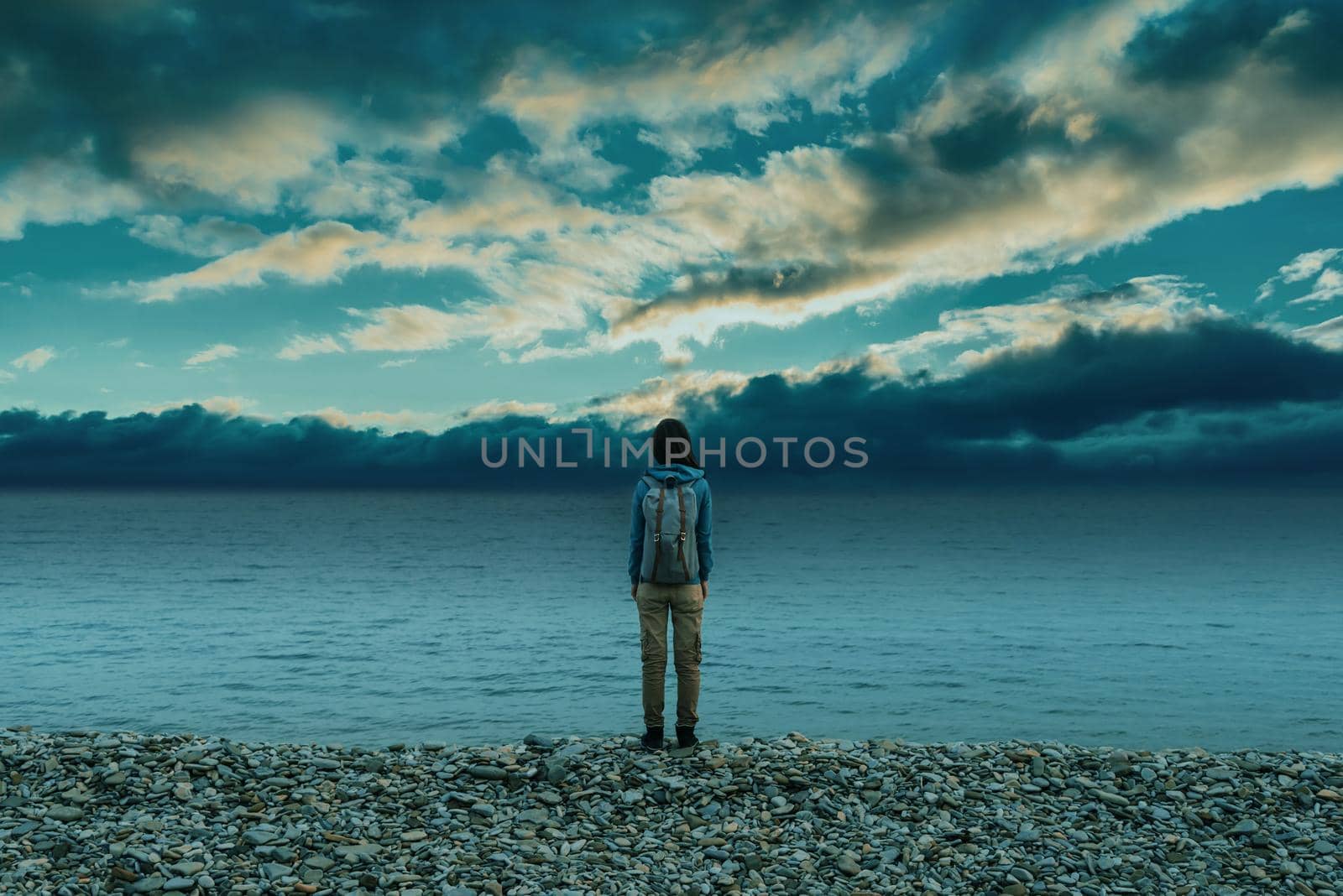 Traveler young woman with backpack standing on pebble coast near the sea and looking at cloudy sky.