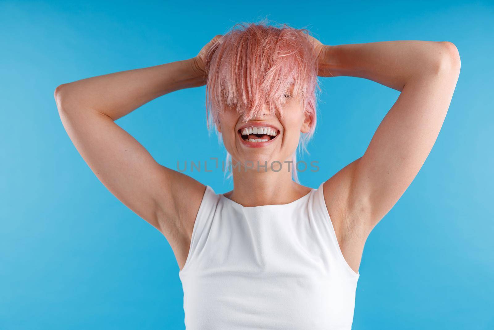 Joyful woman in white shirt playing with smooth straight pink hair ponytail, covering her face with it while posing isolated over blue studio background by Yaroslav_astakhov