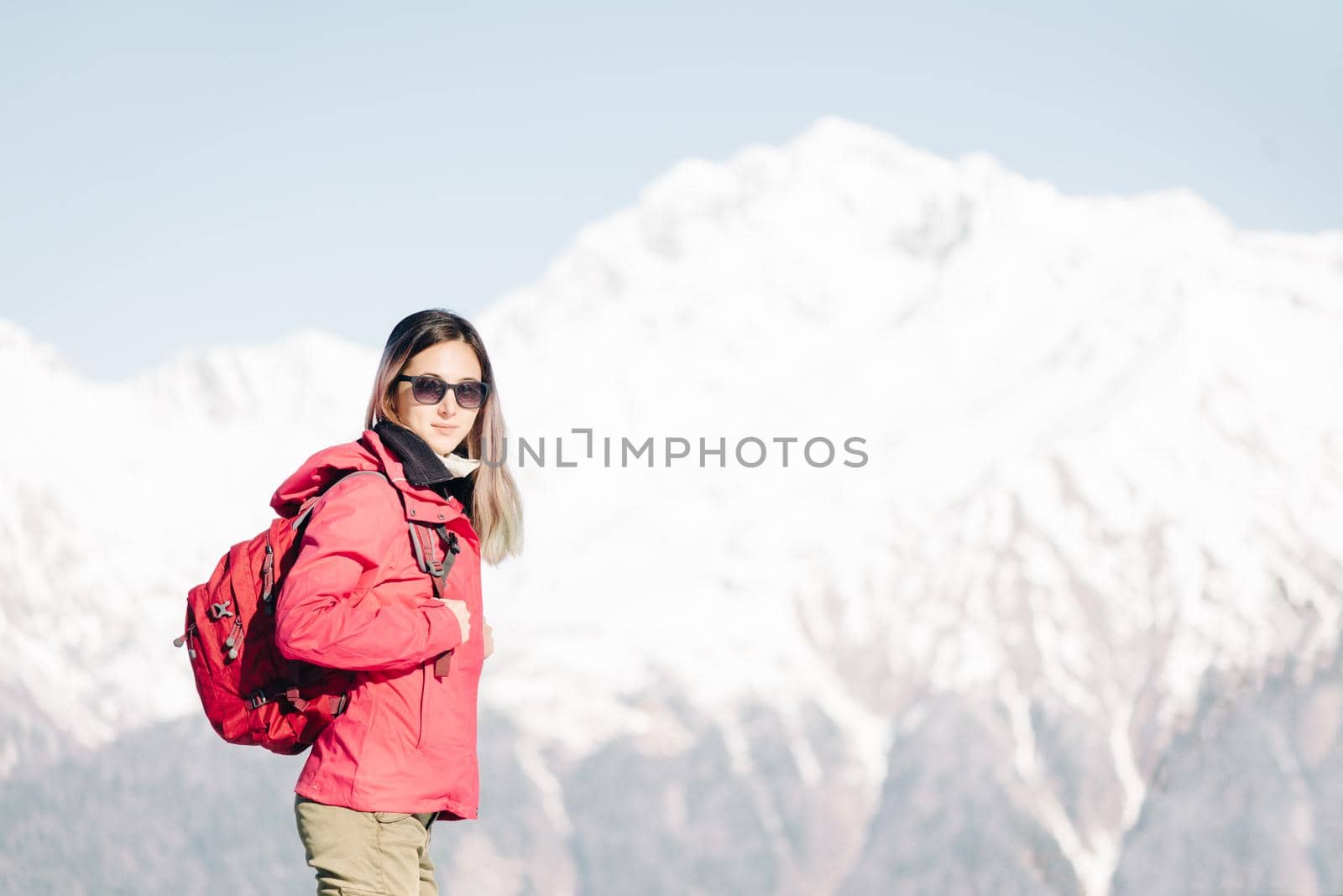 Young woman with backpack standing on background of snowy mountains, looking at camera.