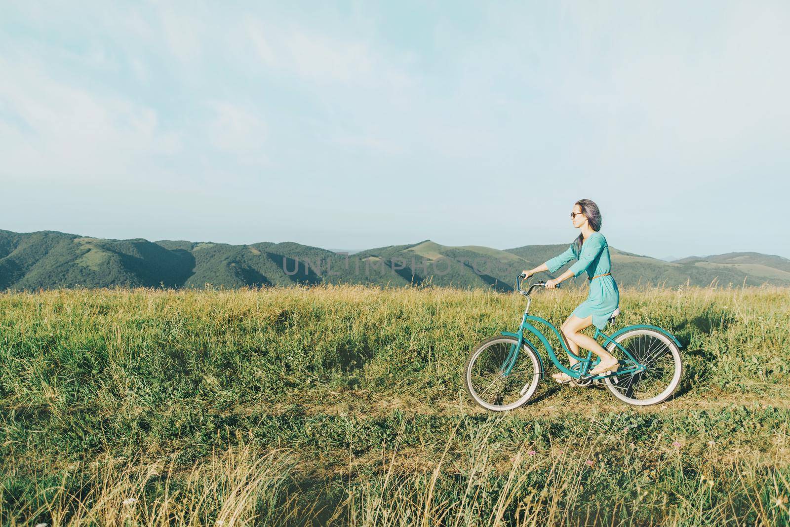 Beautiful young woman wearing in dress riding bicycle on summer meadow, side view.