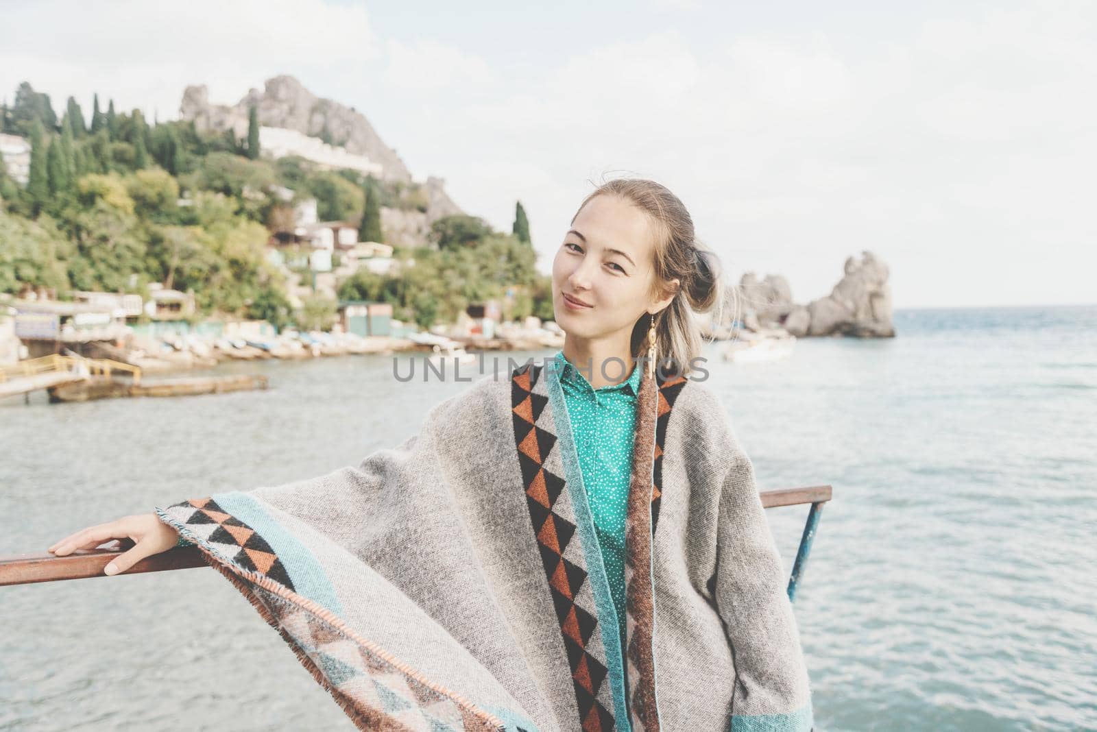 Beautiful smiling young woman wearing in poncho standing on background of sea and coastline, looking at camera.