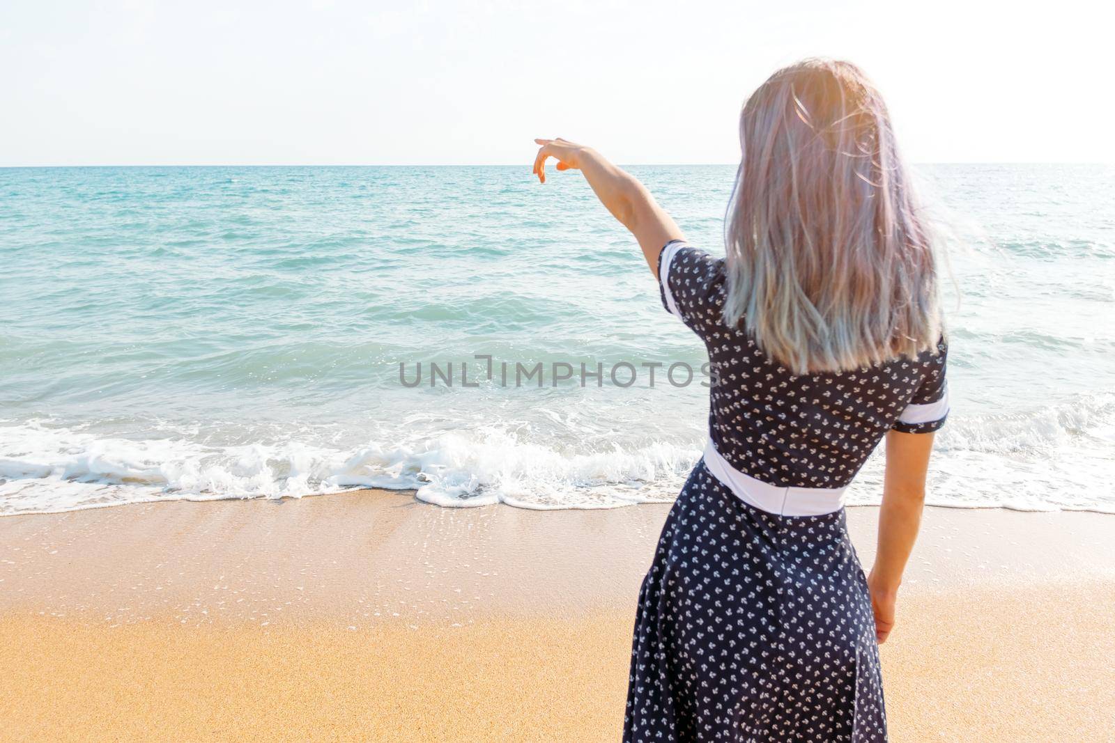 Young woman in dress standing on sand beach and pointing at sea, rear view.