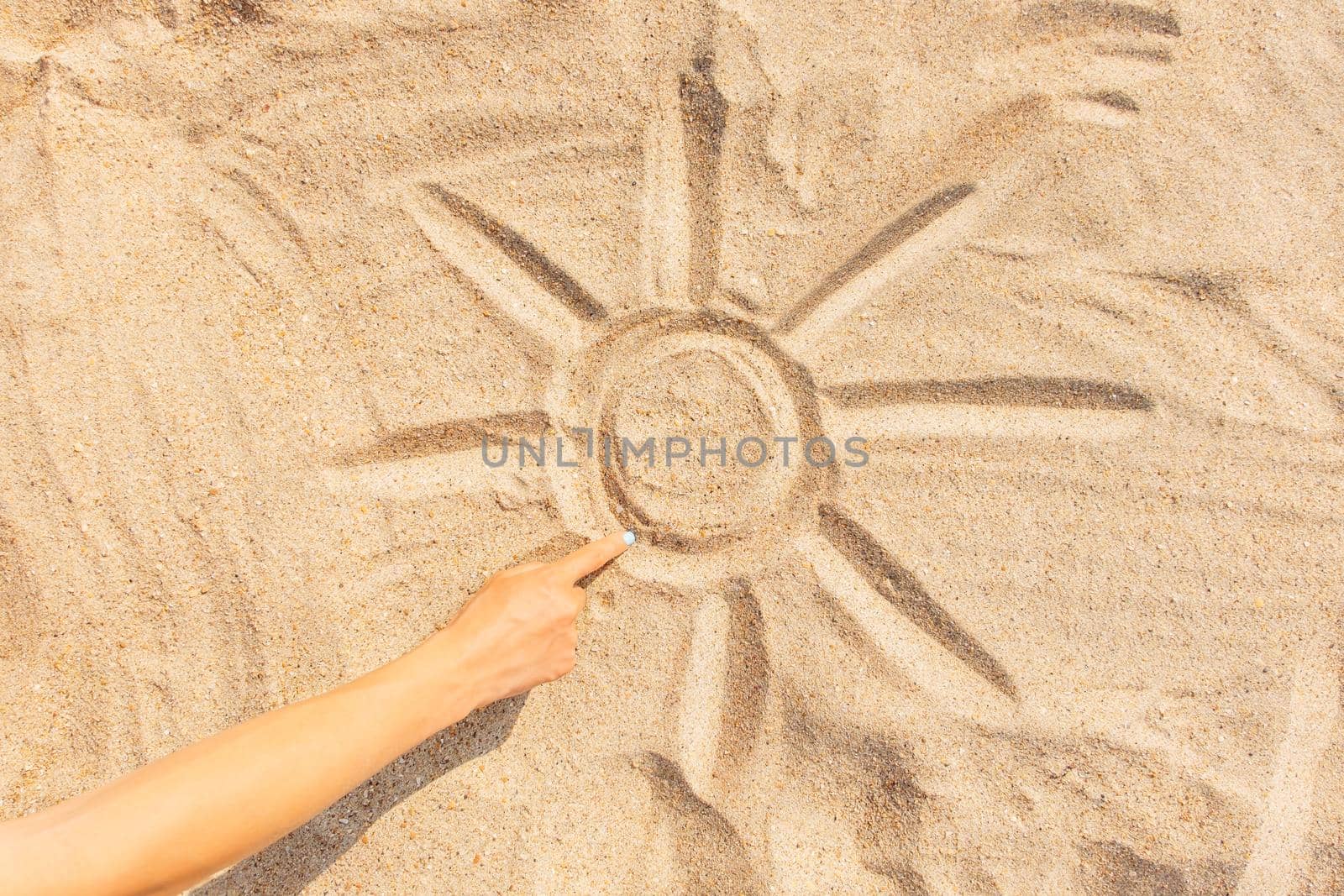 Woman draws a sun with sunbeams on the sand with her finger, view of hand.