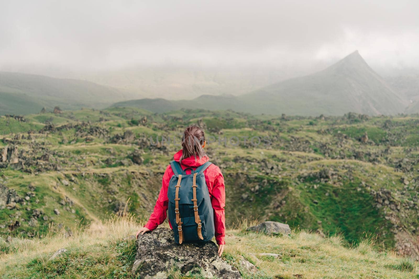 Backpacker young woman sitting on rocky stone and looking at mountains in summer, rear view.