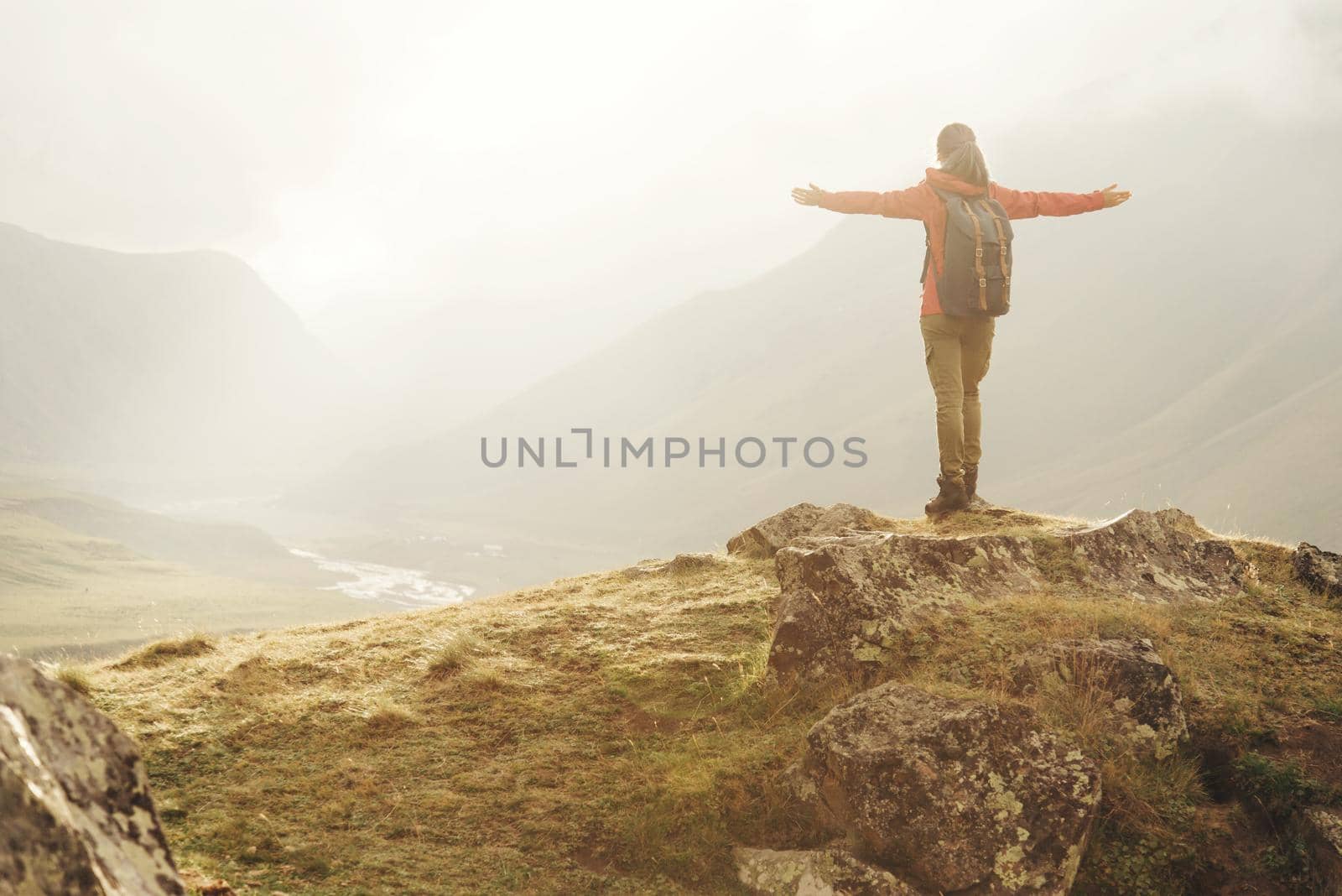 Explorer young woman with backpack standing with raised arms in the mountains at sunset.